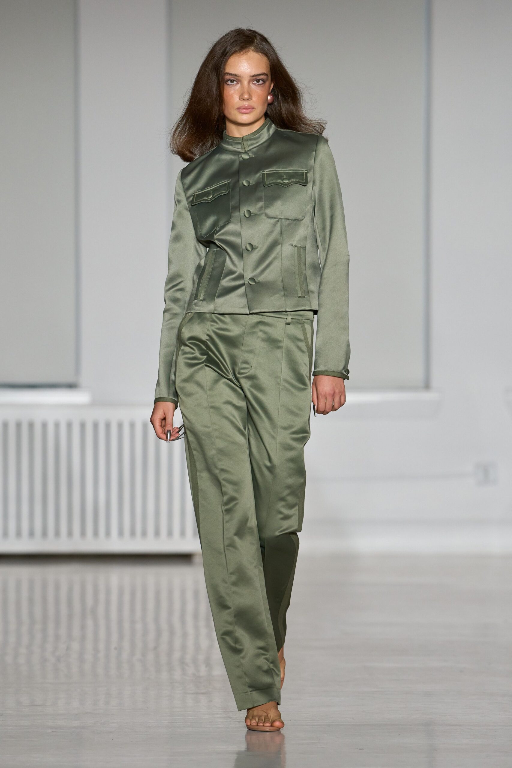 Pink pearl studs at each lobe. Sage satin jacket with a stand collar and piped flap pockets. Matching satin trousers, sharply creased and pooling lightly over bare feet. Walking cool hallway tiles before anyone else at the summer house wakes.