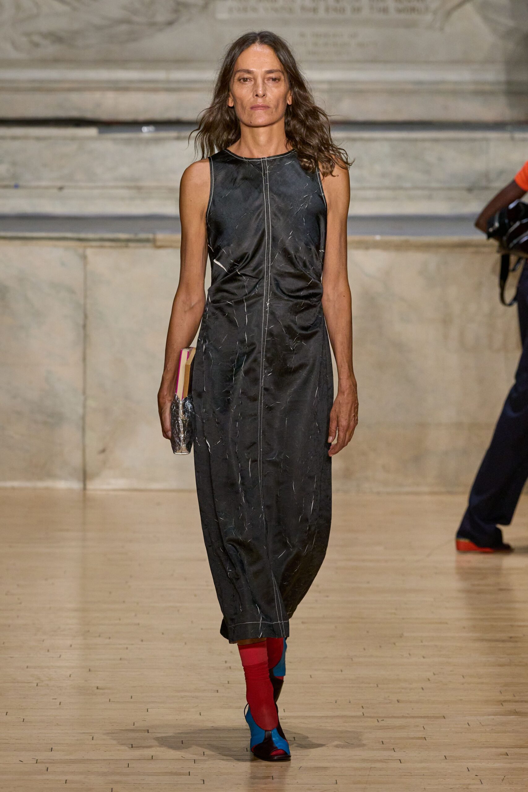Black sleeveless column dress in crinkled satin, raw white seam running dead center. Hardback-shaped clutch wrapped in cloudy plastic, pink page edges flashing. Color-block sock boots in cherry red and pool blue. Leaving a library talk with one borrowed book still under your arm.