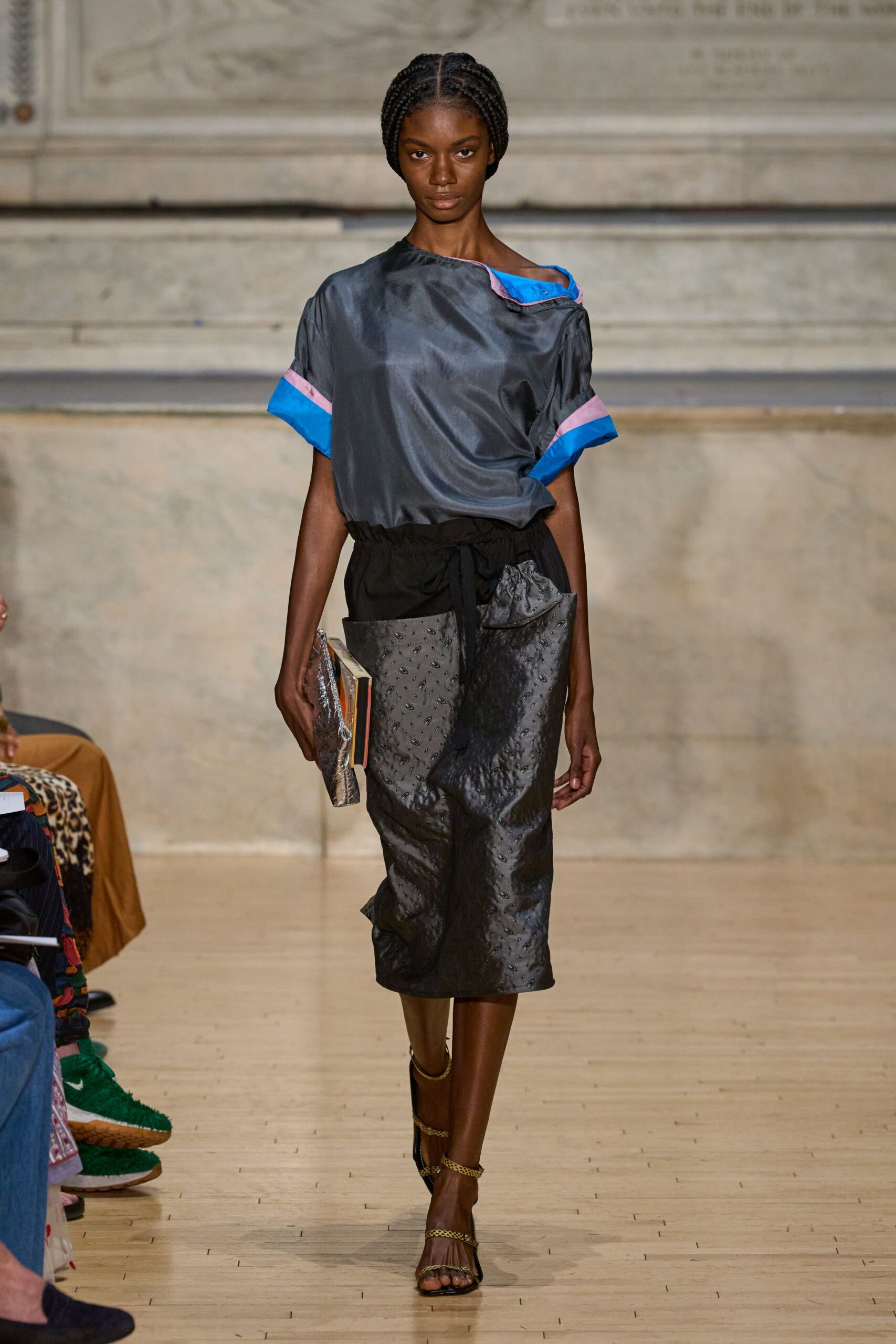 Slate satin top with a skewed neckline and electric-blue turned cuffs. Black midi skirt in puckered embossed satin, paperbag waist tied loosely. Clear plastic-wrapped book carried flat against the thigh. Black sandals with narrow gold chain straps. Leaving the library in rain, one borrowed art book tucked under your arm.