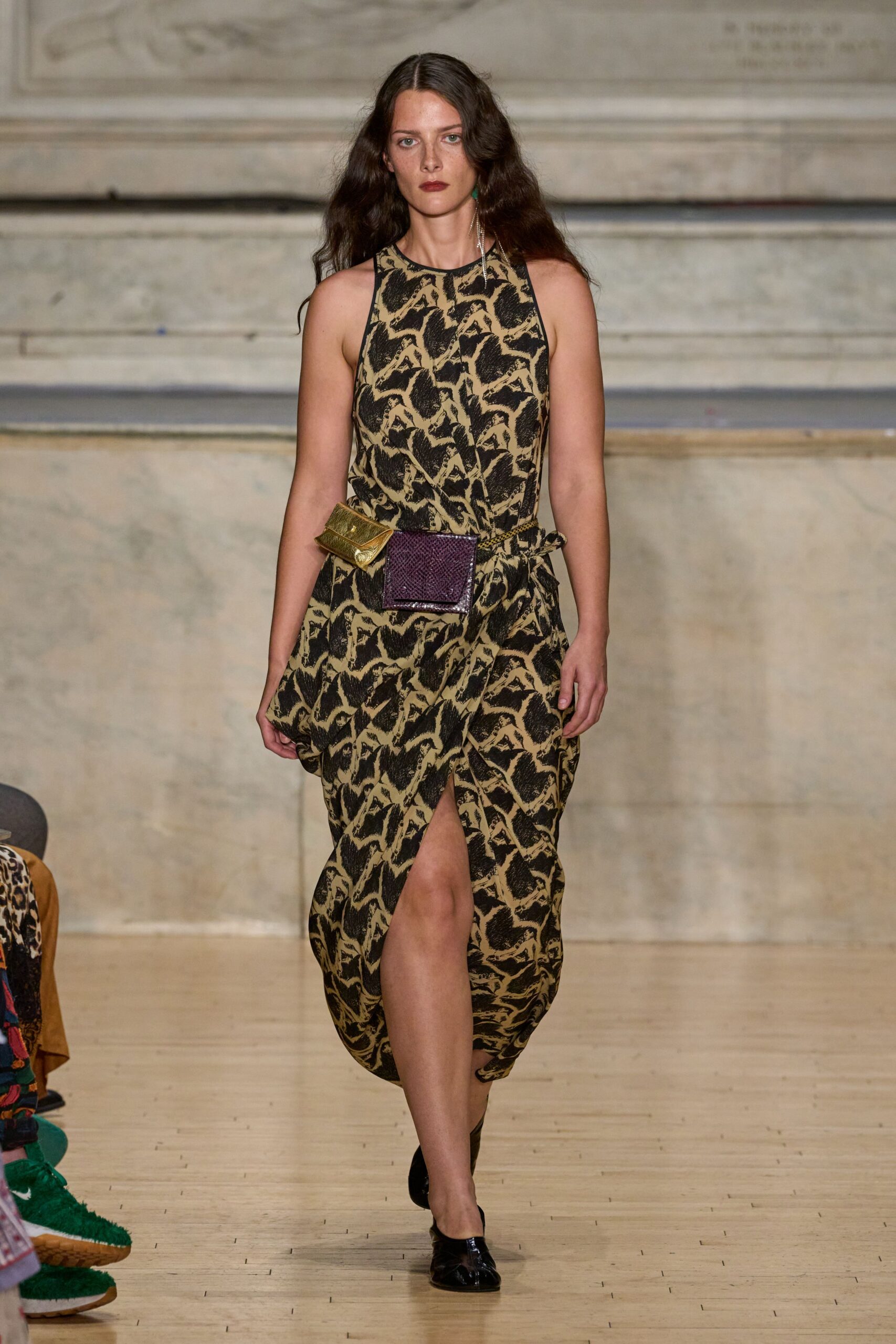 Sand and black print dress with a halter neck and draped slit skirt. Braided waist cord carrying one olive metallic pouch and one plum snakeskin box. Black patent flats, sharply pointed. Walking through a civic hall after the heat has broken.