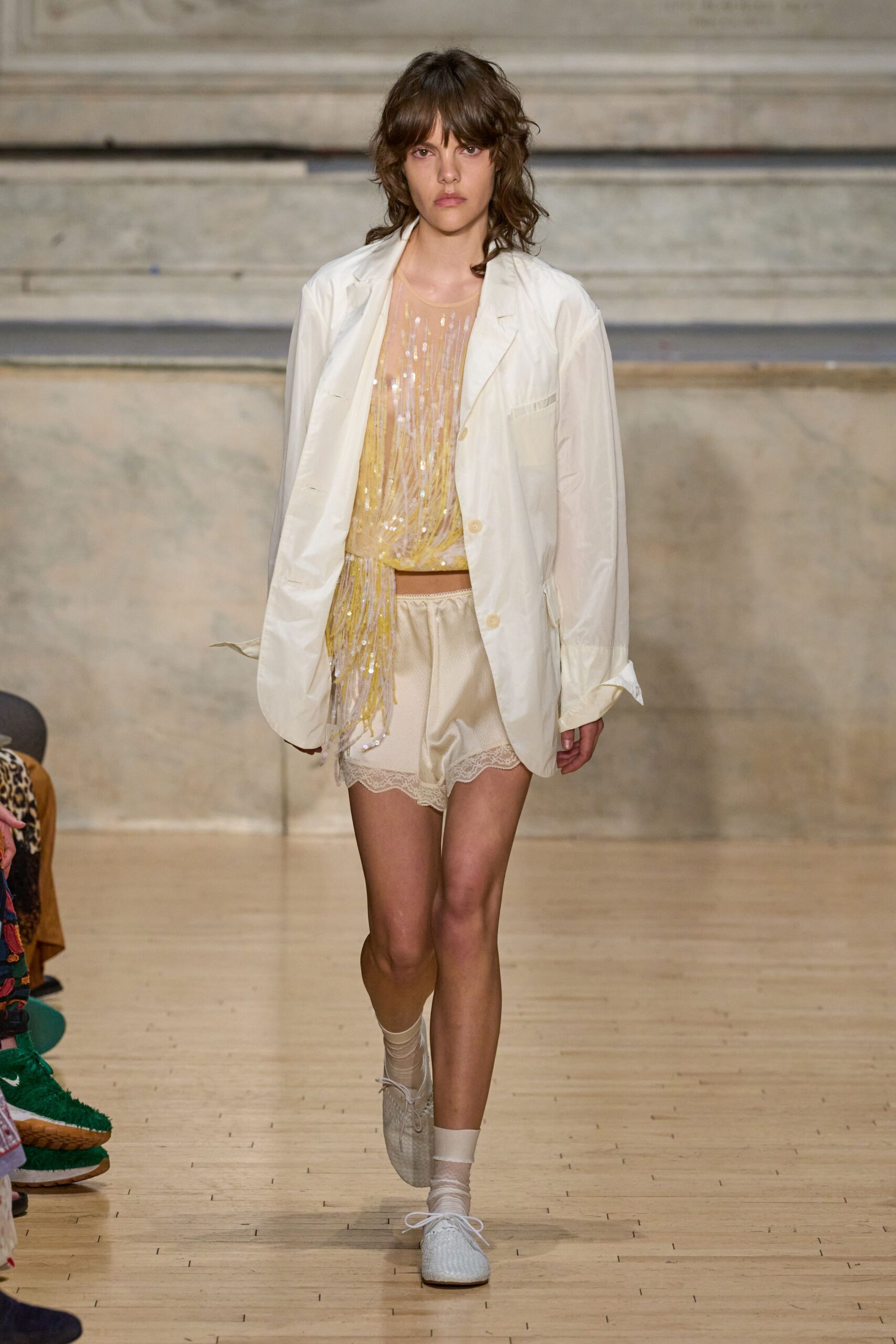 Oversized ivory blazer in papery cloth, cuffs left rumpled open. Pale yellow beaded fringe top over sheer nude mesh. Champagne tap shorts with a scalloped lace hem. Sheer ankle socks, slightly slouched. White woven lace-up flats. Morning after a wedding, crossing the hotel lobby for ice.