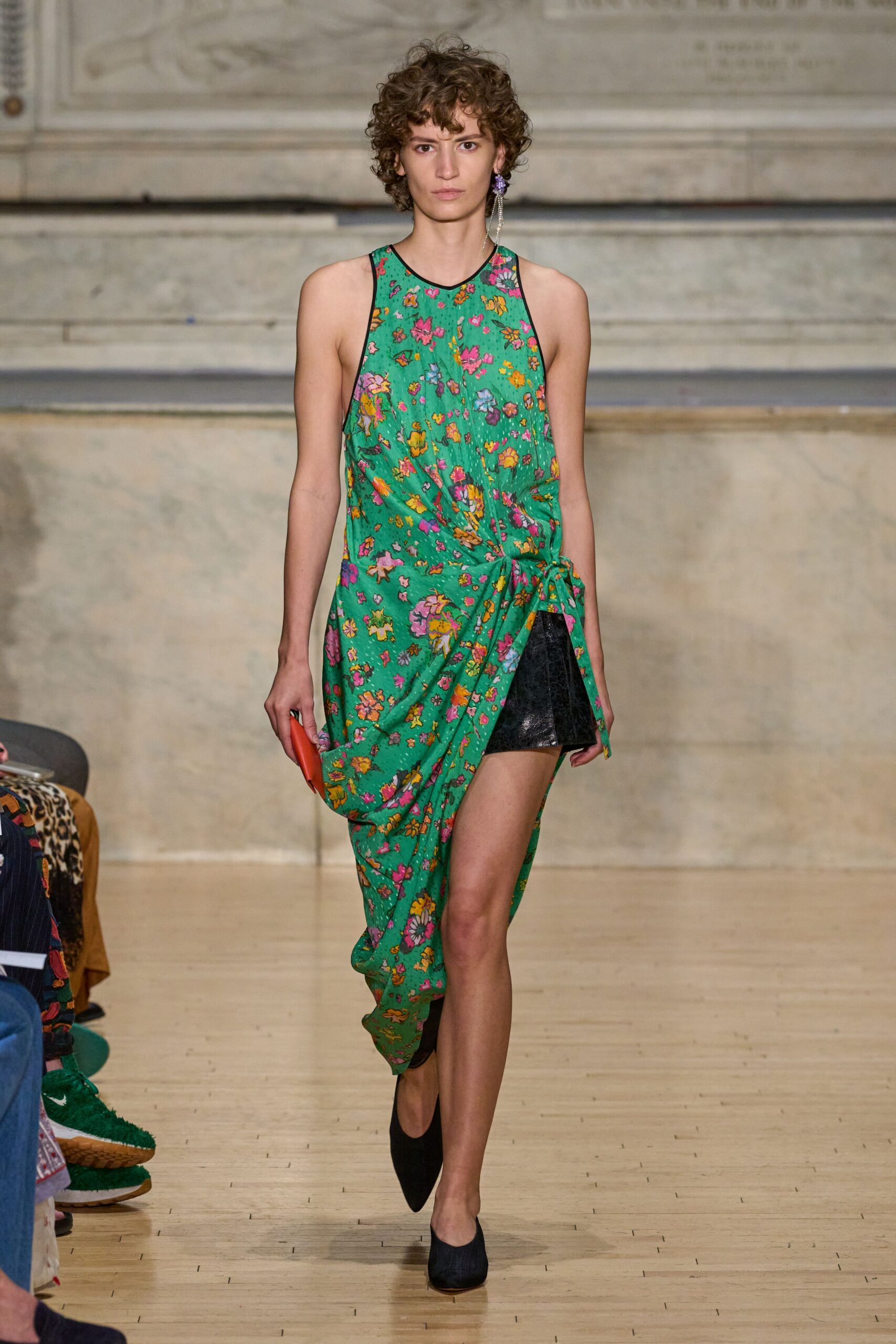 Lavender cluster earring with one thin crystal strand brushing the shoulder. Emerald floral slip dress draped and knotted low at one hip. Black crackle-finish mini peeking from the split underlayer. Slim coral clutch in the hand. Black almond-toe flats. Crossing a museum atrium with a folded note you have not opened yet.
