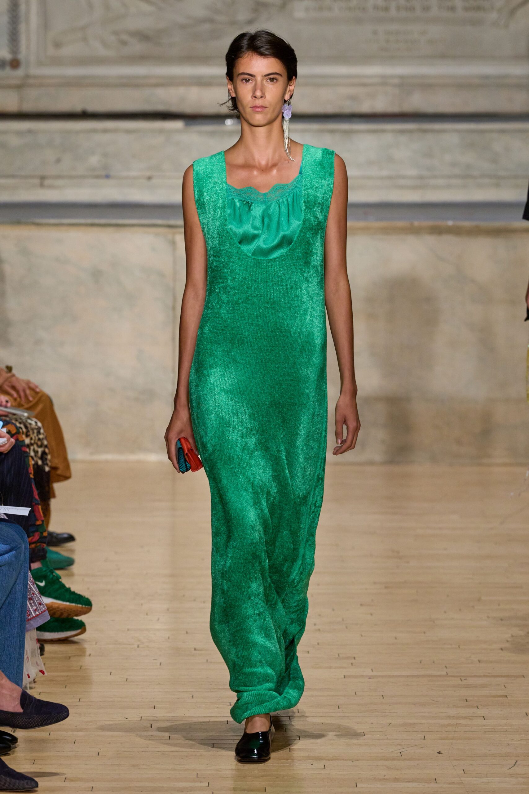 Sea-green brushed knit column dress with a satin bib inset and scalloped edge. Lilac flower drop with a curtain of rhinestone strands. Small red clutch with a blue textured side panel. Black patent flats, squared off and mirror-bright. Crossing a museum atrium after the wedding, still carrying the favors.