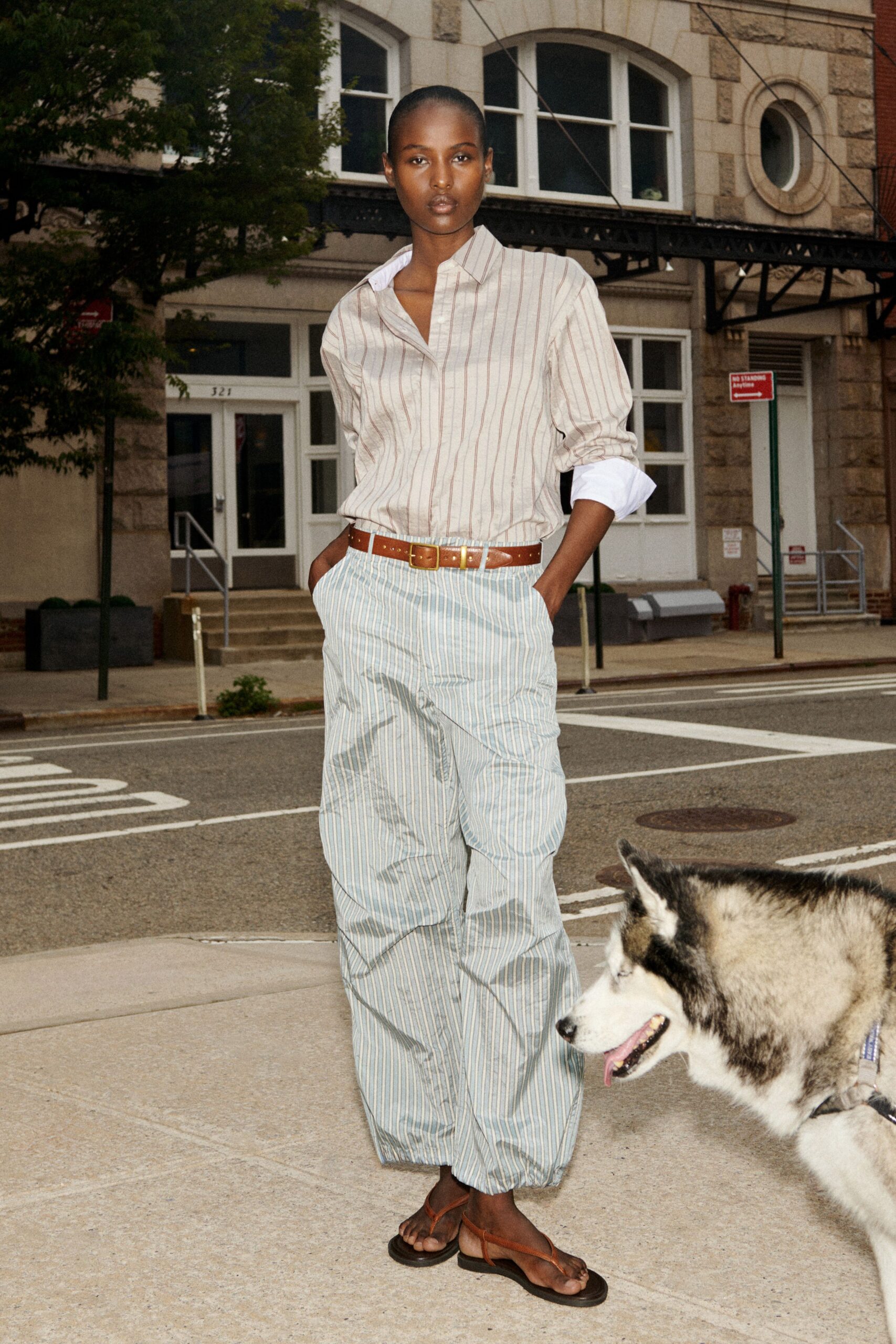 Cream striped cotton shirt, white cuffs turned back below the forearm. Tan leather belt with a square brass buckle. Powder-blue striped trousers, ballooning softly into gathered hems. Brown leather toe-post sandals on thick black soles. Crossing for coffee while the husky pulls toward the park.