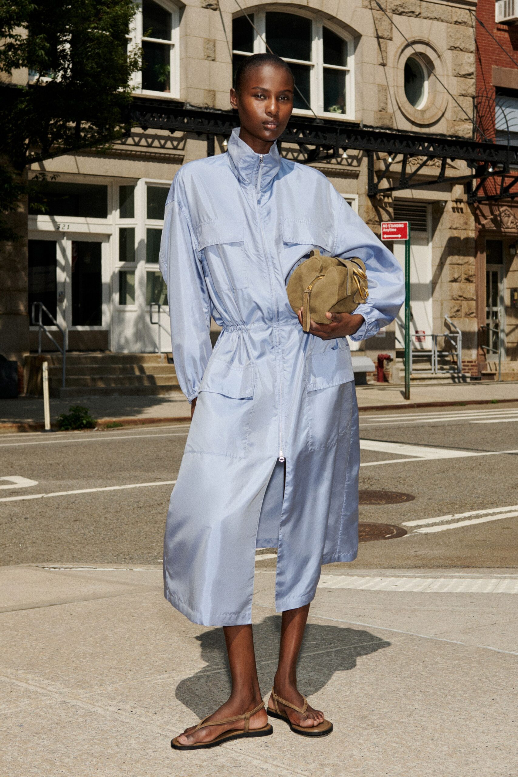 Powder-blue nylon coat dress, high zip collar and drawstring waist. Olive suede pouch tucked under the arm, brass zip pull hanging. Flat tan leather thong sandals with slim wraparound straps. Crossing to the corner deli before the heat lifts off the pavement.