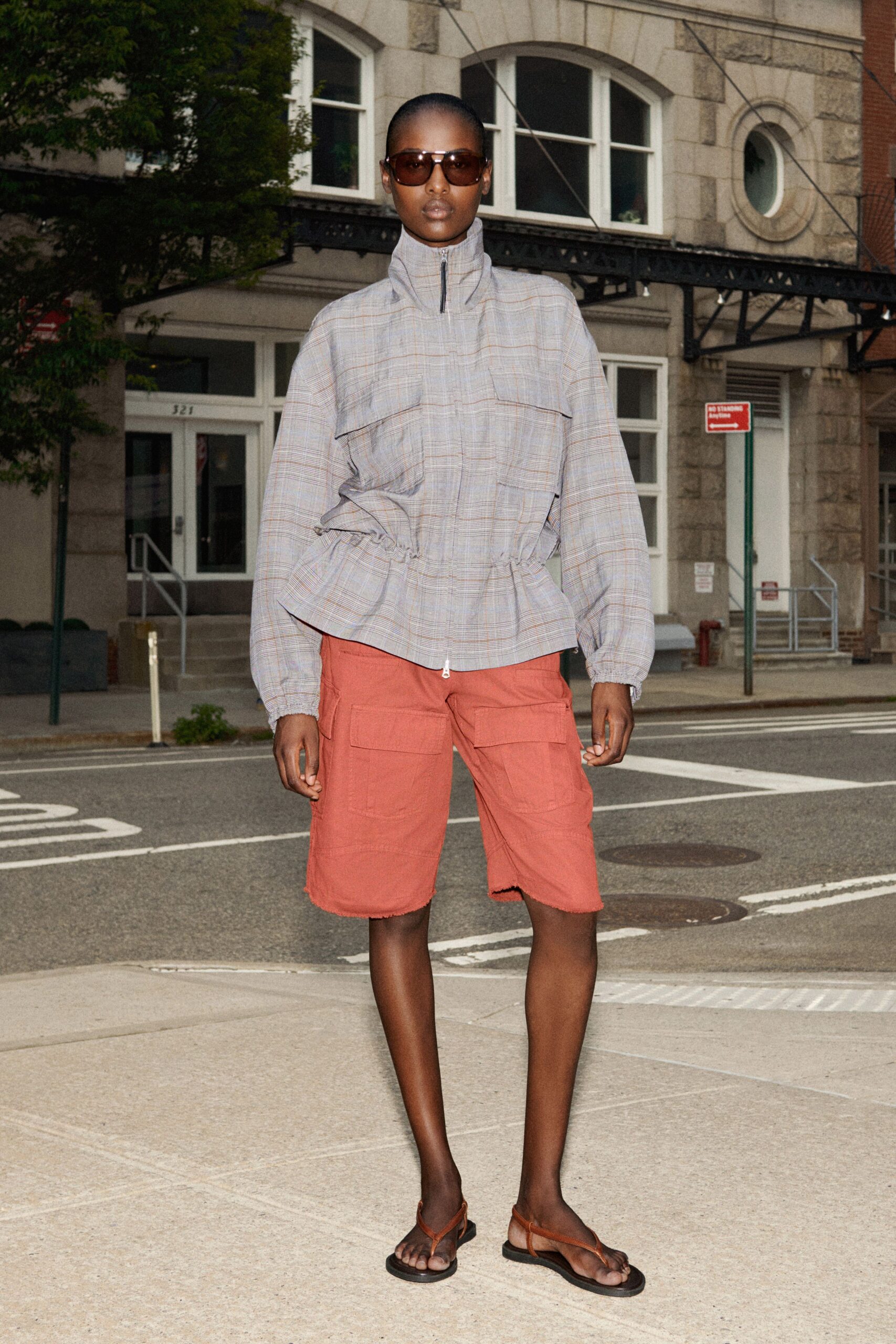 Brown gradient shield lenses with a slim silver bridge. Pale glen-plaid anorak, funnel zip neck and drawstring peplum waist. Faded clay cargo shorts with raw hems and broad flap pockets. Brown leather thong sandals on black rubber soles. Crossing side streets just after rain, takeaway coffee cooling too fast.