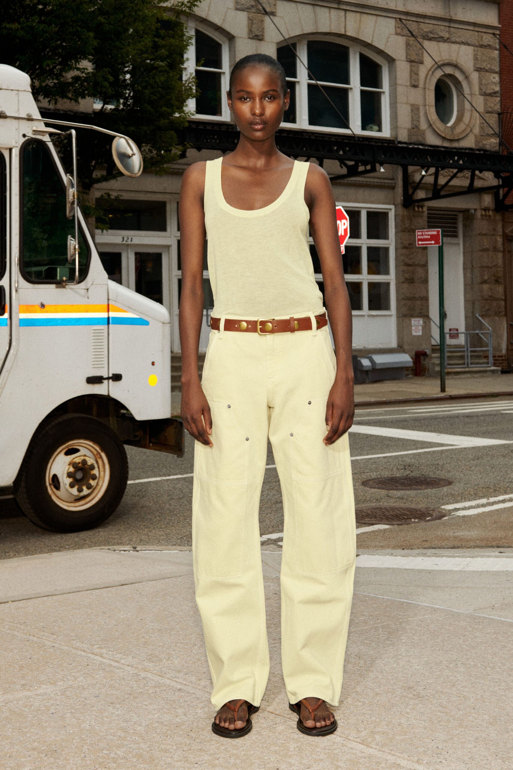 Pale butter scoop-neck tank in thin jersey, nearly translucent at the waist. Brown leather belt with oversized brass studs and a square buckle. Cream wide-leg trousers with curved knee seams and tiny metal rivets. Brown leather thong sandals, flat black soles. Crossing to the corner deli before the heat lifts off the pavement.