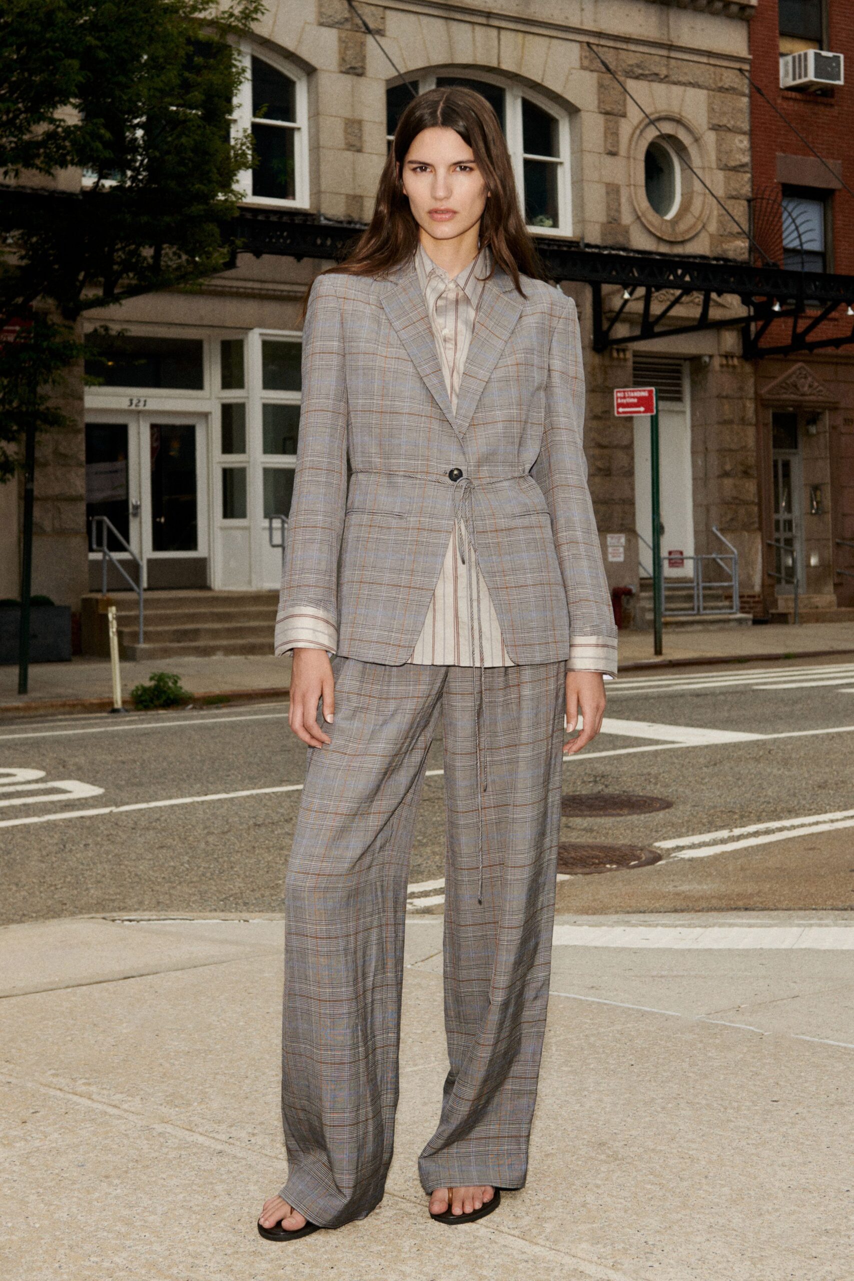 Grey prince-of-wales blazer cinched with a dangling self-tie through one button. Pale striped shirt, long cuffs turned past the jacket sleeves. Matching wide trousers breaking heavily over flat thong sandals. Black thong sandals, toes fully exposed. Crossing to the corner deli before the office air-conditioning wakes up.