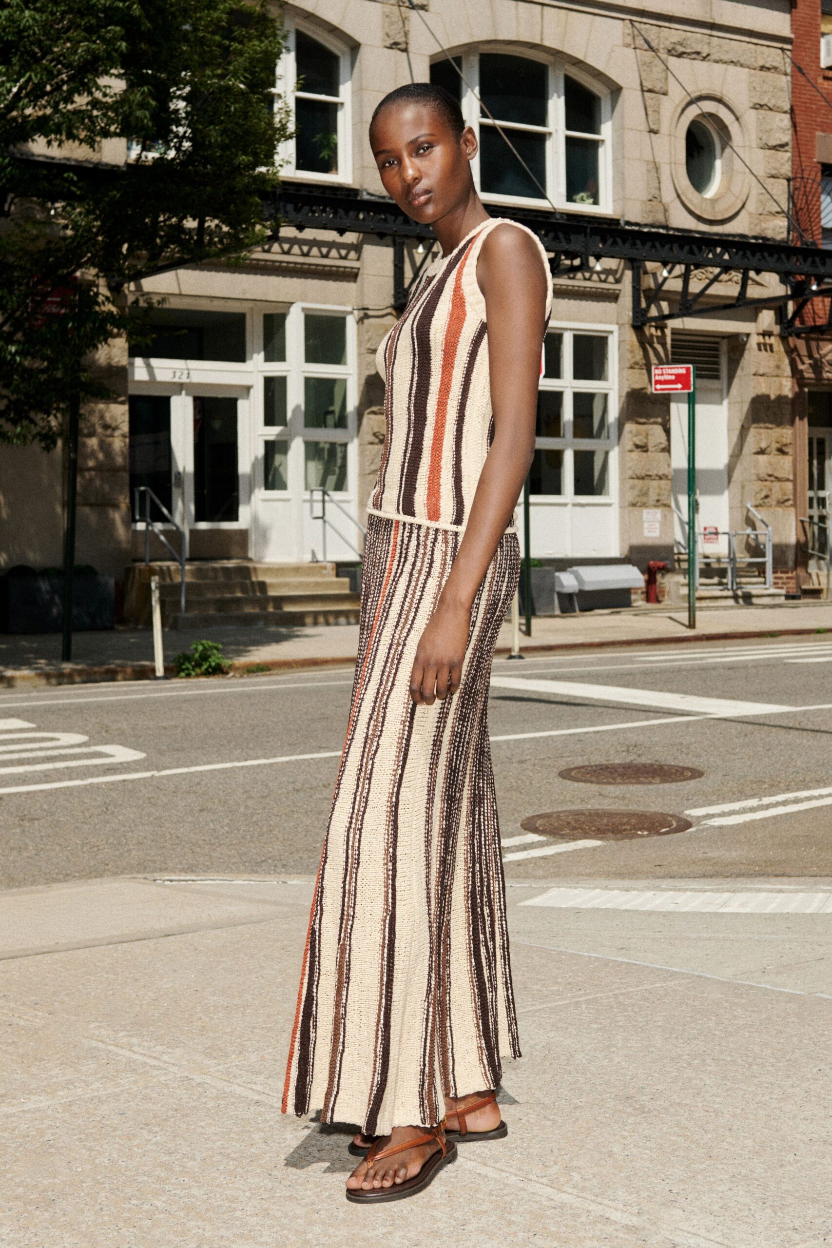 Open crochet tank in cream with rust and espresso vertical stripes. Matching crochet maxi, softly flared with a narrow slit breaking the hem. Brown leather thong sandals wrapping once at the ankle. Crossing a quiet downtown block before the heat lifts off the pavement.