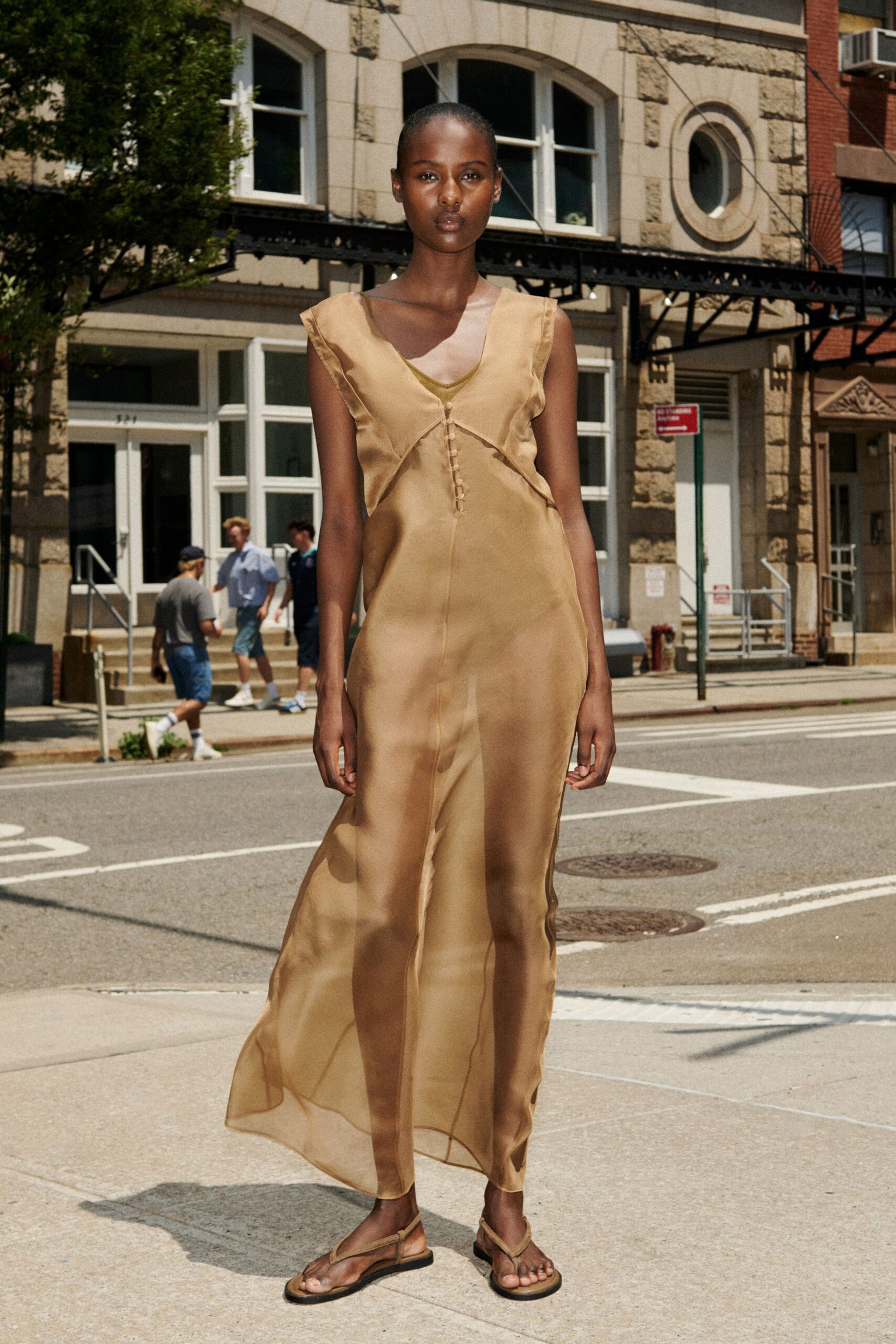 Sheer camel organza column dress over a slip, tiny covered buttons at the sternum. Flat thong sandals with slim matching straps. Crossing to the deli before noon heat turns the avenue metallic.