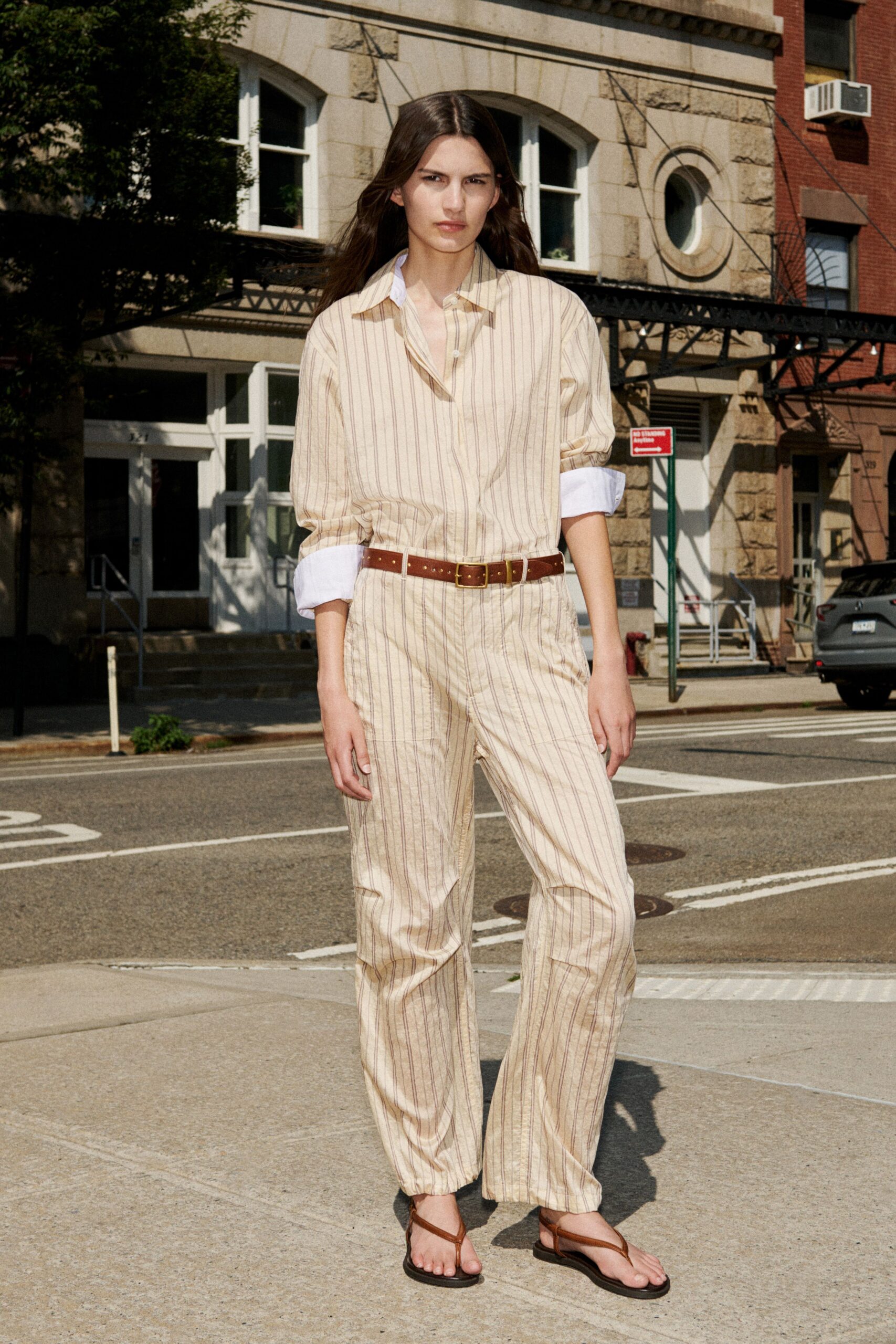 Cream striped cotton shirt, white cuffs turned back thick at the wrist. Matching drawstring trousers, soft creases pooling lightly over the sandal. Chestnut leather belt with a small brass buckle. Brown leather thong sandals on flat black soles. Crossing a quiet block at noon before the heat starts to stick.