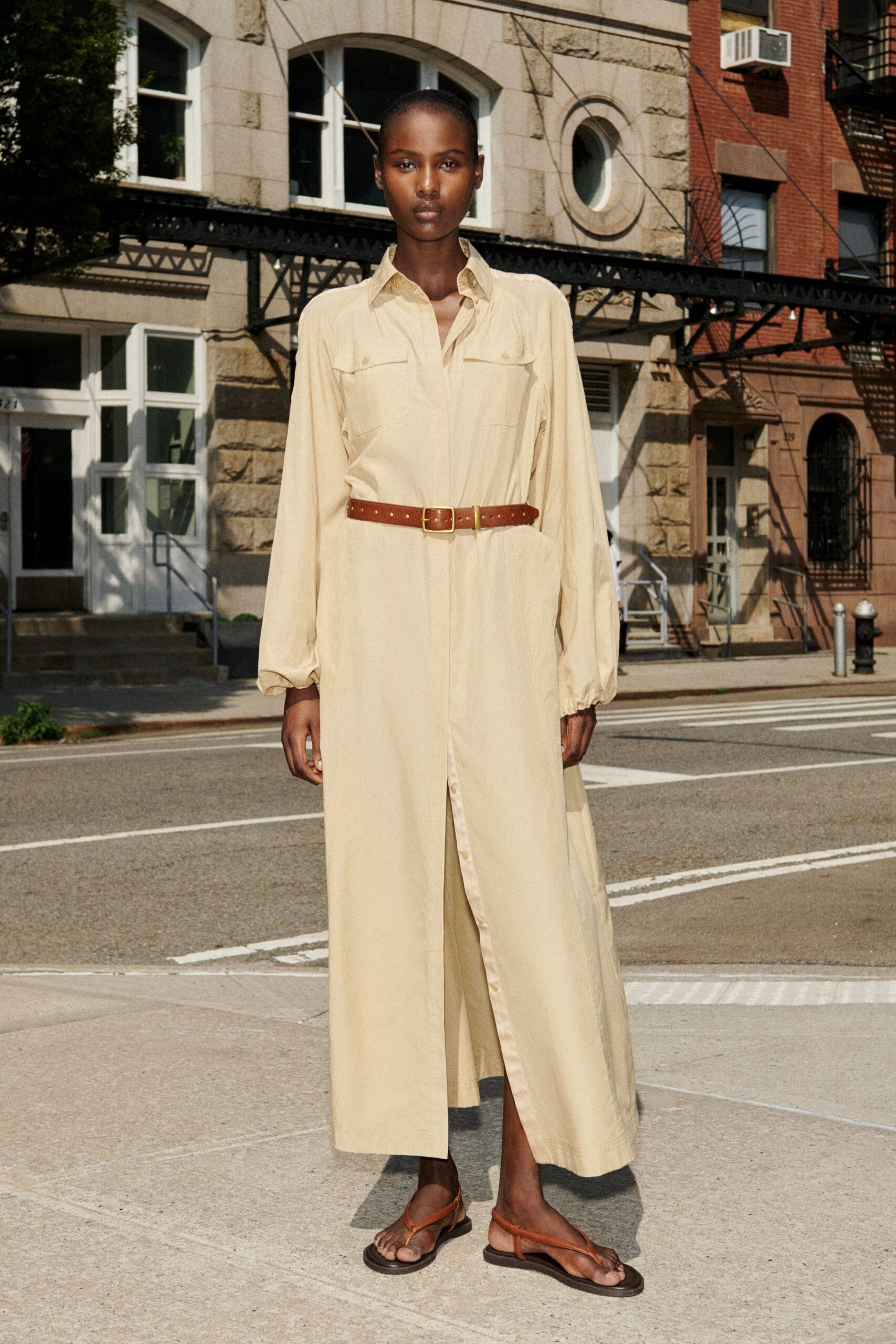 Buttercream shirt dress in fluid twill, flap pockets and a low button placket. Chestnut leather belt, punched close with a square brass buckle. Brown leather thong sandals on flat black soles. Crossing a sunstruck side street before the bodega shuts for ice.