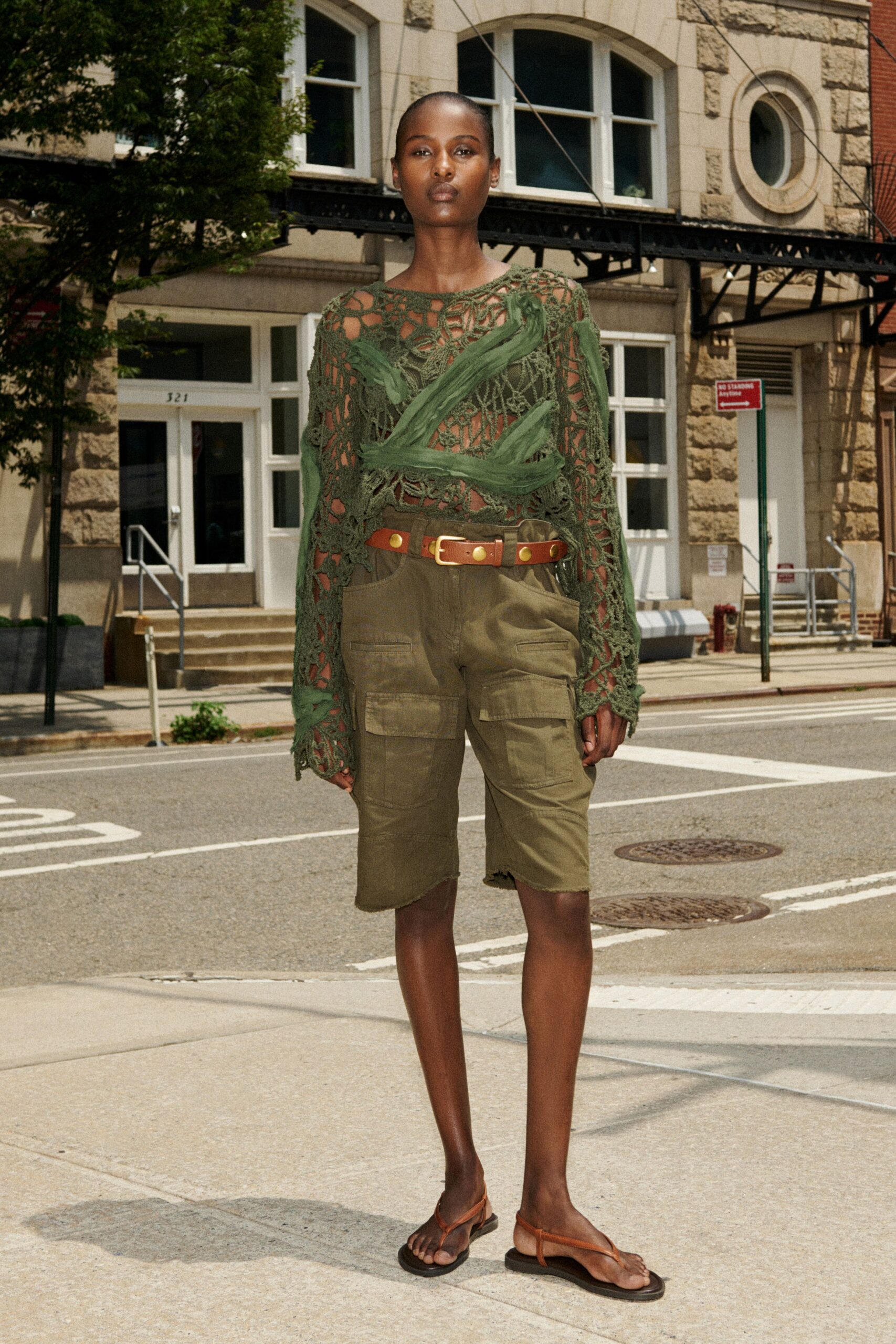 Olive crochet sweater, shredded net traced with gauzy bands across the chest. Tan leather belt with oversized brass grommets and square buckle. Olive canvas cargo shorts, flap pockets and a raw uneven hem. Flat brown leather thong sandals. Crossing downtown before noon, asphalt already throwing heat back up.
