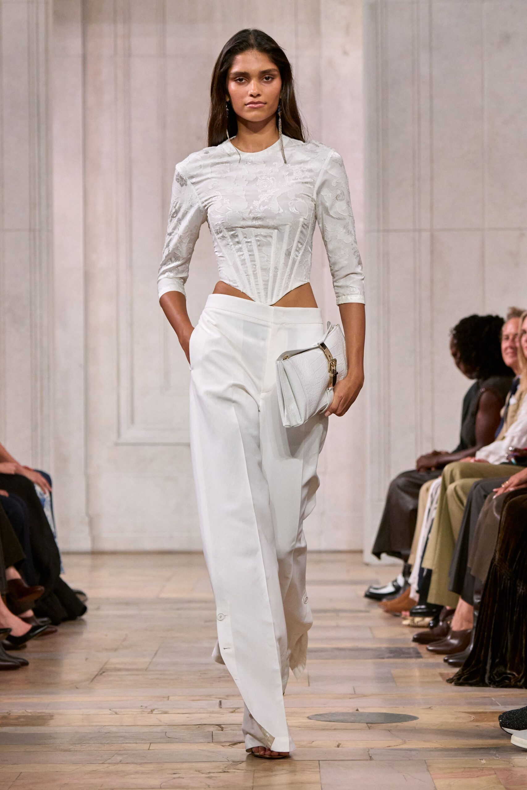 Pearl-white brocade top with corset boning tapering to a sharp point. Ivory wide-leg trousers, pressed creases and buttoned ankle tabs grazing the floor. White crocodile-embossed clutch with a gold corner clasp. Long crystal drop earrings. For crossing a hotel lobby before noon with yesterday still intact.