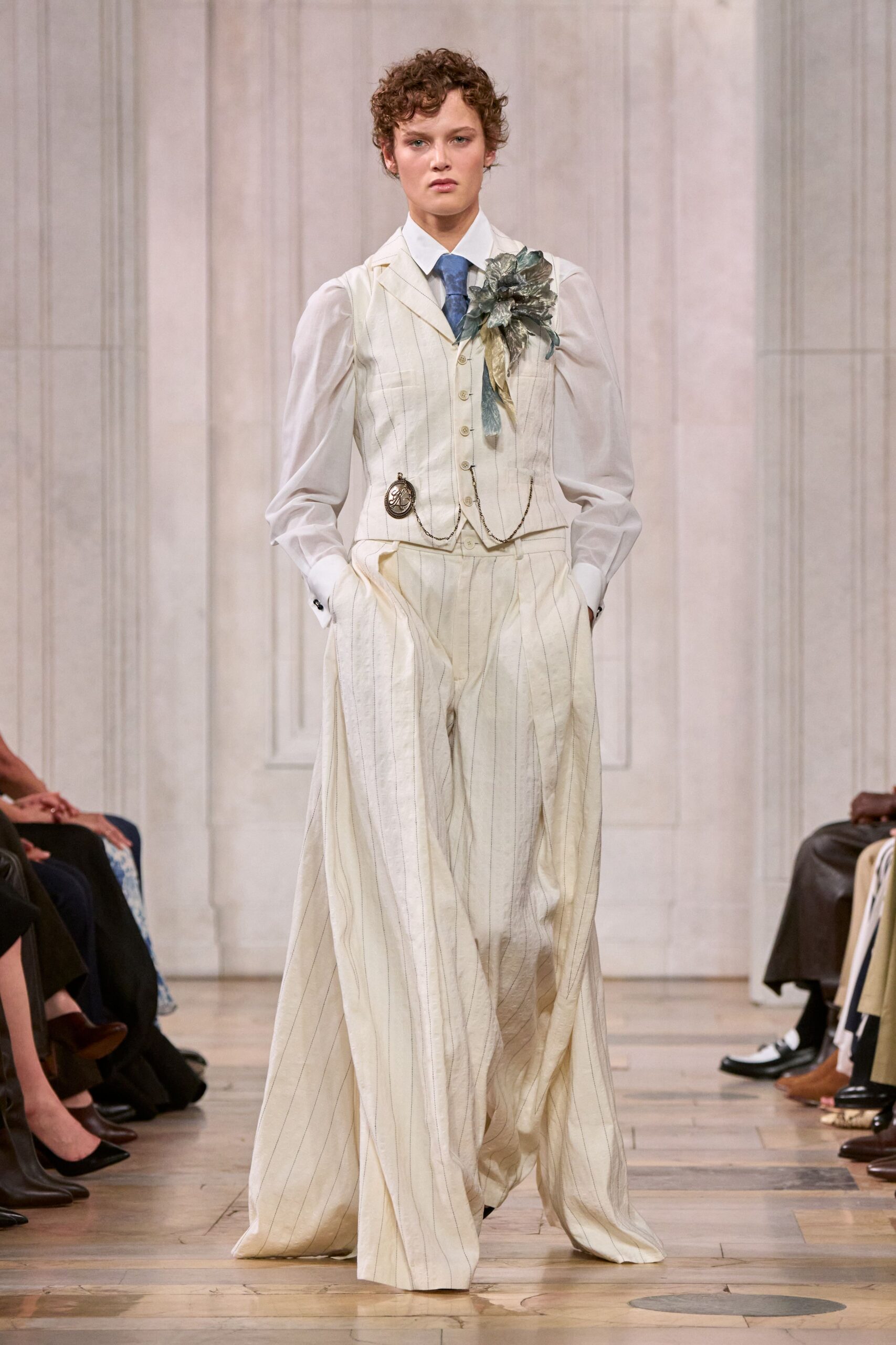 White shirt with sheer bishop sleeves and a sharp collar. Dusty blue tie knotted tight at the throat. Ivory pinstriped waistcoat with tiny buttons, watch chain, oversized silk flower. Matching wide trousers falling in soft pooled folds. For reading old letters in a cold house after the estate sale.