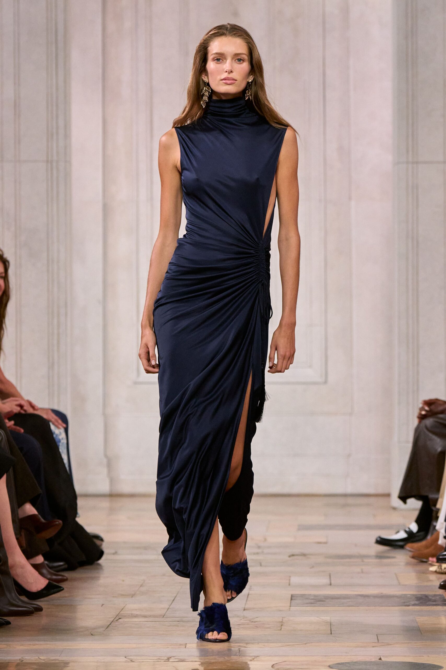 Ink-navy jersey gown with a rolled turtleneck, side cutout and ruched hip tie. Antique-gold chandelier drops brushing the jaw. Navy feather-trim mules. For slipping into a hotel bar after the wedding band has packed up.