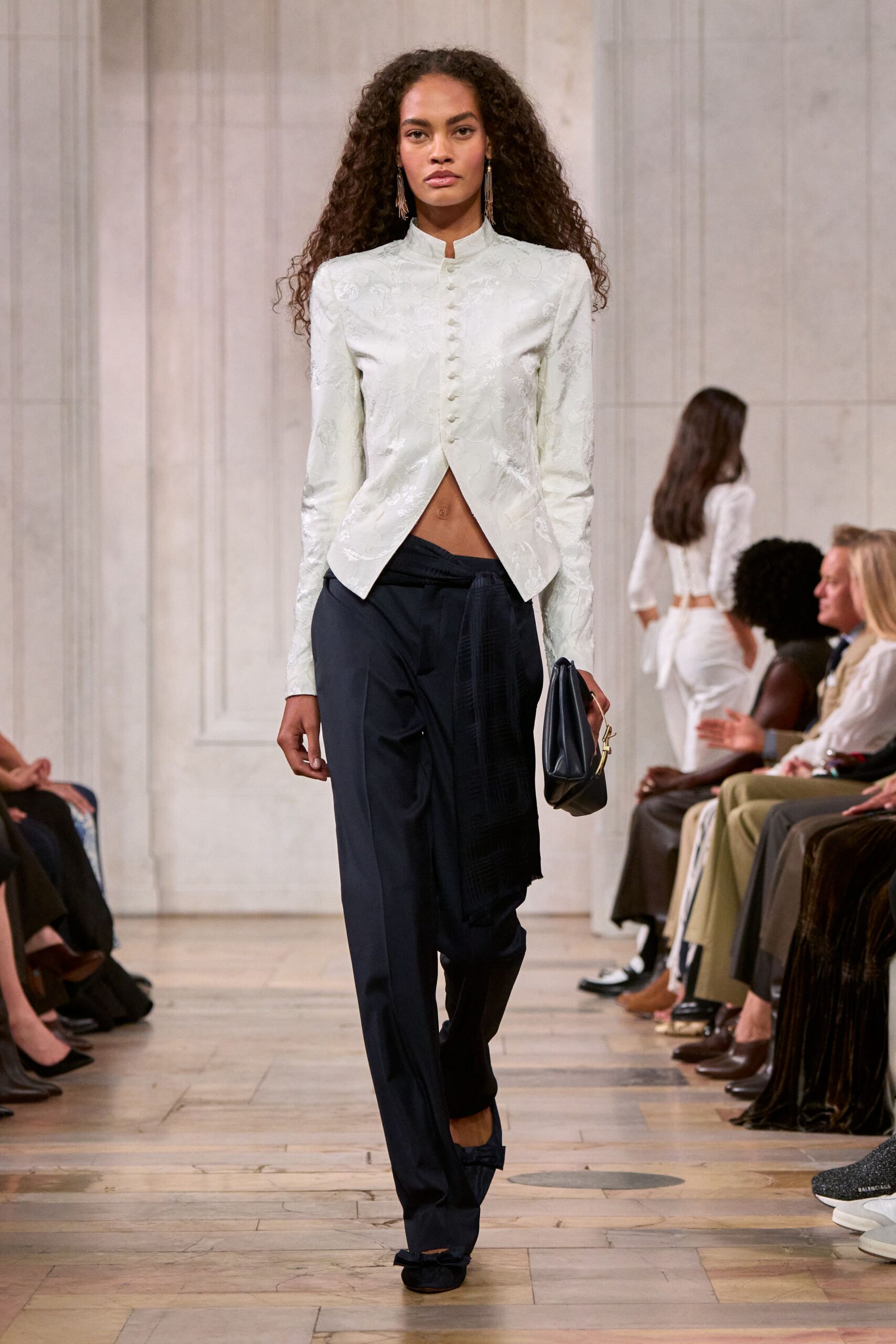 Ivory floral brocade jacket with a stand collar and covered buttons. Ink tailored trousers with a long scarf panel knotted off the waistband. Soft black foldover clutch with a small gold side clasp. Black satin flats tied in bows across the vamp. Crossing a hushed hotel corridor before the wedding guests spill out.