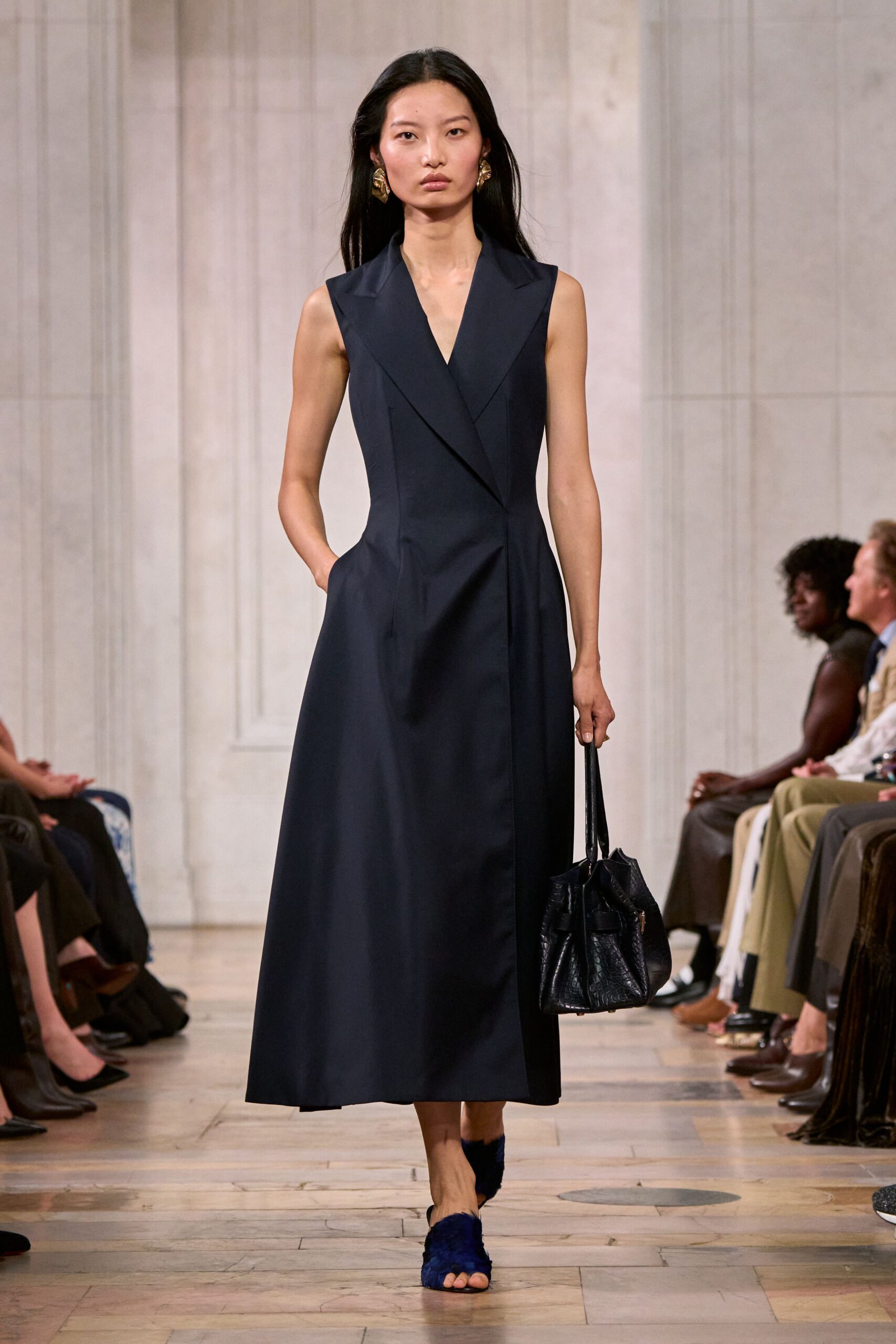 Crumpled gold earrings, petal-shaped against the jaw. Ink-dark sleeveless coat dress with broad lapels and a wrapped front seam. Black croc-embossed bucket bag collapsing softly at the sides. Midnight fur sandals with bare toes peeking through. Walking into a hotel lobby before noon, carrying nothing but the room key.