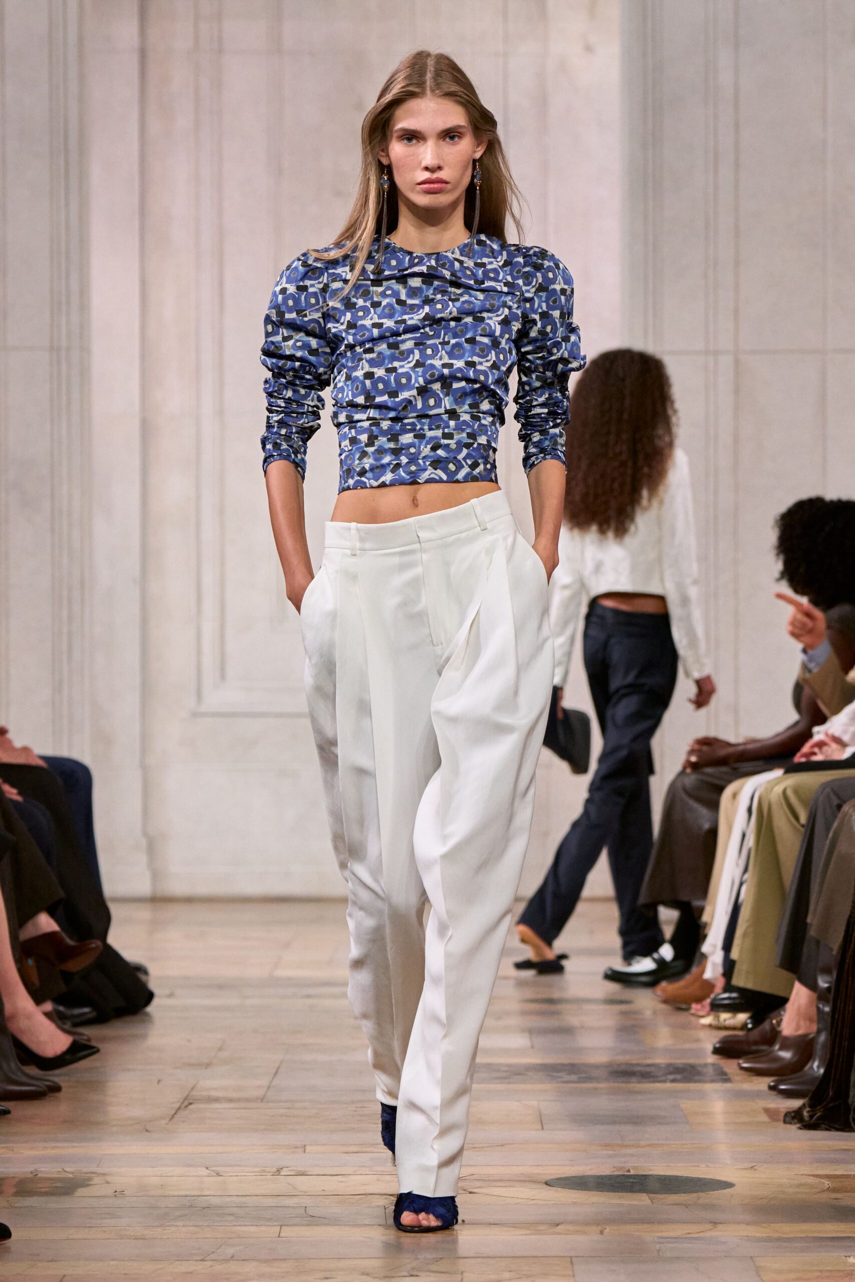 Blue geometric printed top, ruched across the body with a skewed collar. White tailored trousers with deep pleats and slanted hip pockets. Long drop earrings brushing the shoulders. Navy feathered peep-toe heels. Walking into a city wedding after the rain finally let up.
