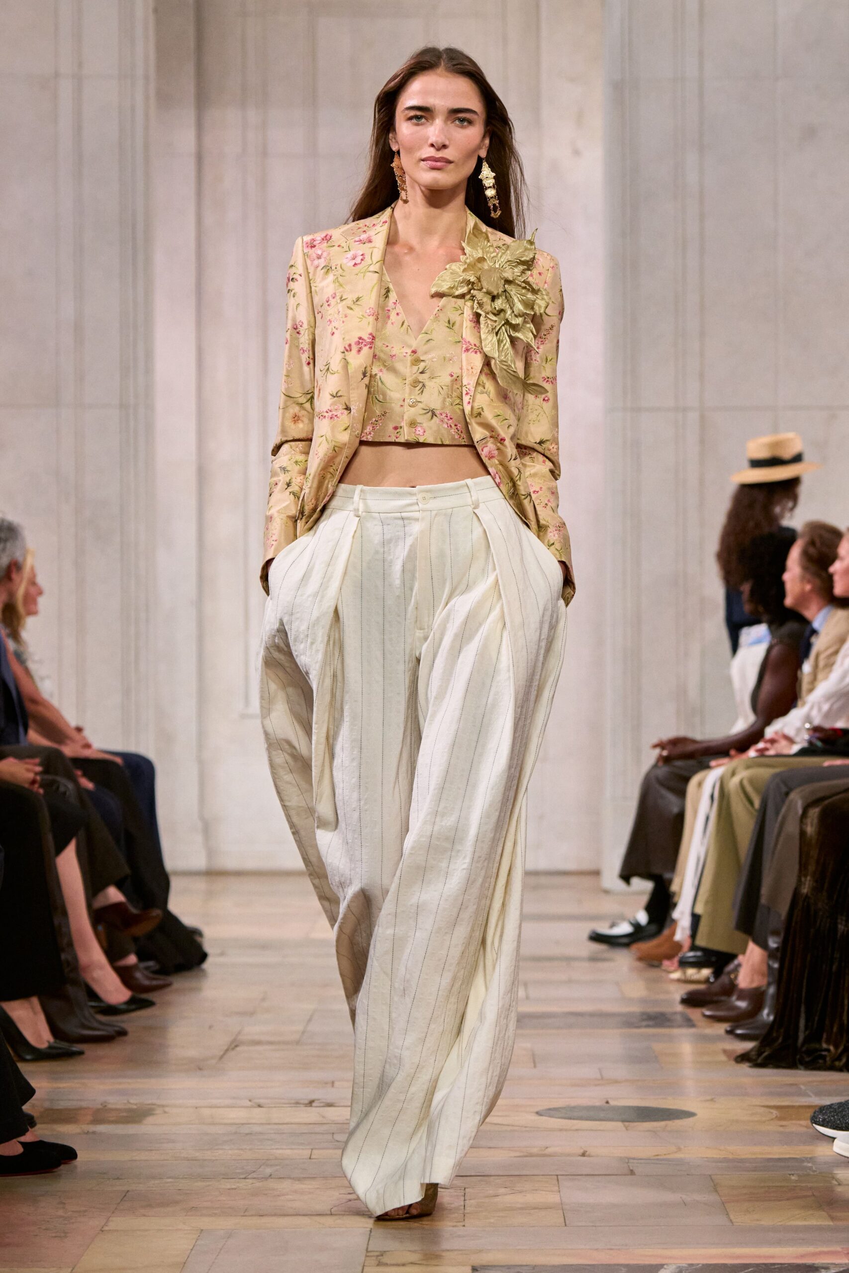 Long baroque drop earrings in hammered gold. Champagne floral brocade blazer with a crumpled flower at one lapel. Matching cropped waistcoat, tiny pearl buttons down the front. Ivory pinstriped trousers cut very full through the pleats. Tan peep-toe heels. Garden wedding reception, one shoe already sinking into the lawn.