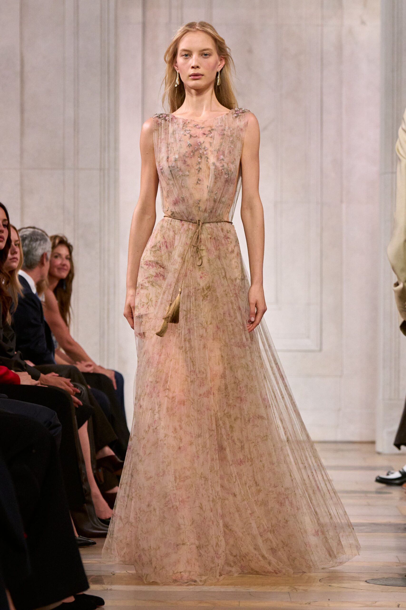 Blush tulle gown printed with faded meadow flowers, softly pleated throughout. Twisted cord belt knotted low, tassel ends brushing the hip. Long drop earrings in pale metallic shards. Crossing a hotel ballroom after the string quartet has packed away.