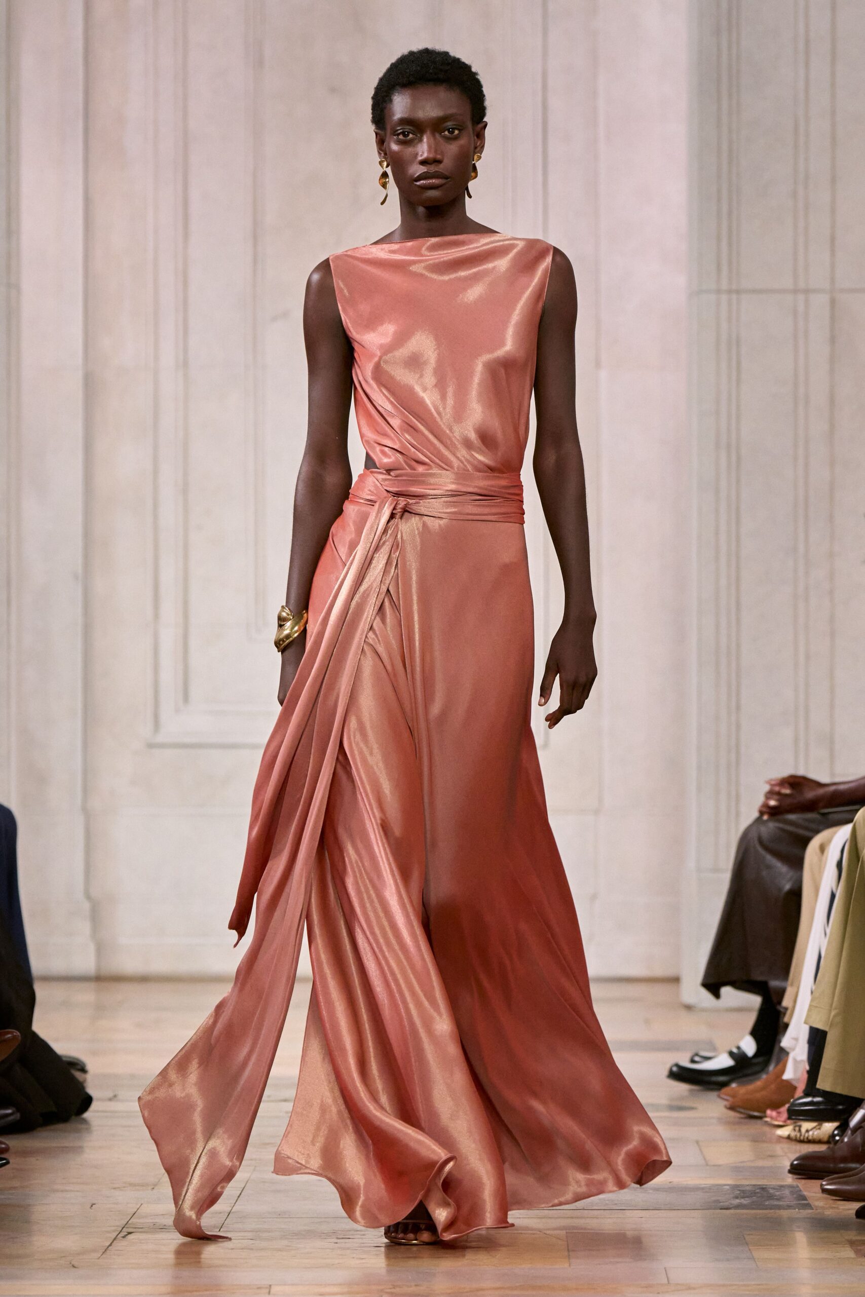 Rose-copper satin gown with a bateau neck and sash-wrapped hip. Gold drop earrings, irregular and molten-looking. Wide gold cuff, crumpled like folded foil. Barely there metallic sandals under the sweeping hem. Crossing a hotel ballroom after midnight, one ribbon end still skimming the floor.