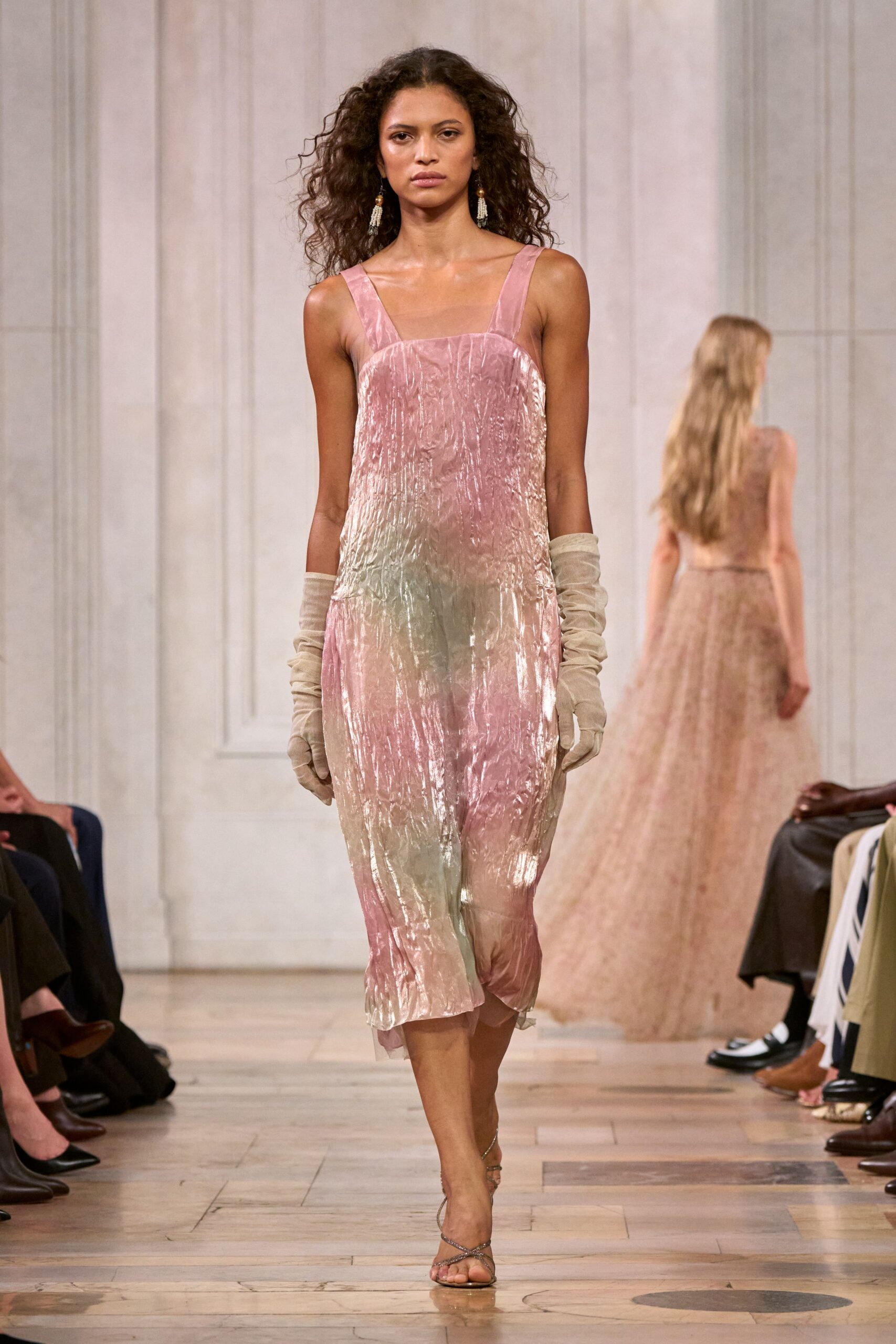 Crushed velvet slip dress in blush and sage, hem wavering unevenly. Oat suede opera gloves ruched down the forearms. Long tassel earrings with a bead at the lobe. Silver wrap sandals, barely there across the toes. Crossing a hotel ballroom after the flowers have started to wilt.