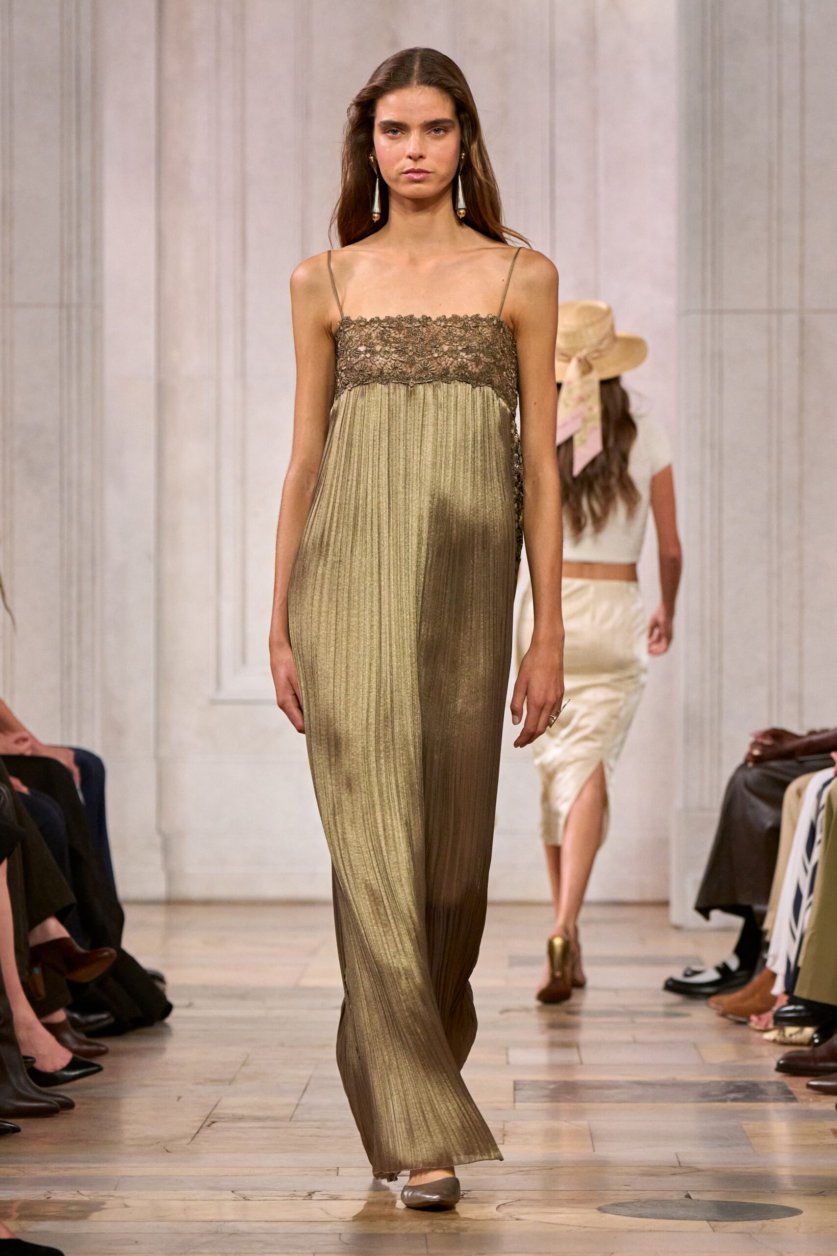 Olive-gold plisse column dress with a scalloped lace camisole yoke. Long drop earrings with pale tassels and bead tips. Taupe satin flats, softly squared at the toe. Walking across a hotel lobby just after the string quartet packed up.