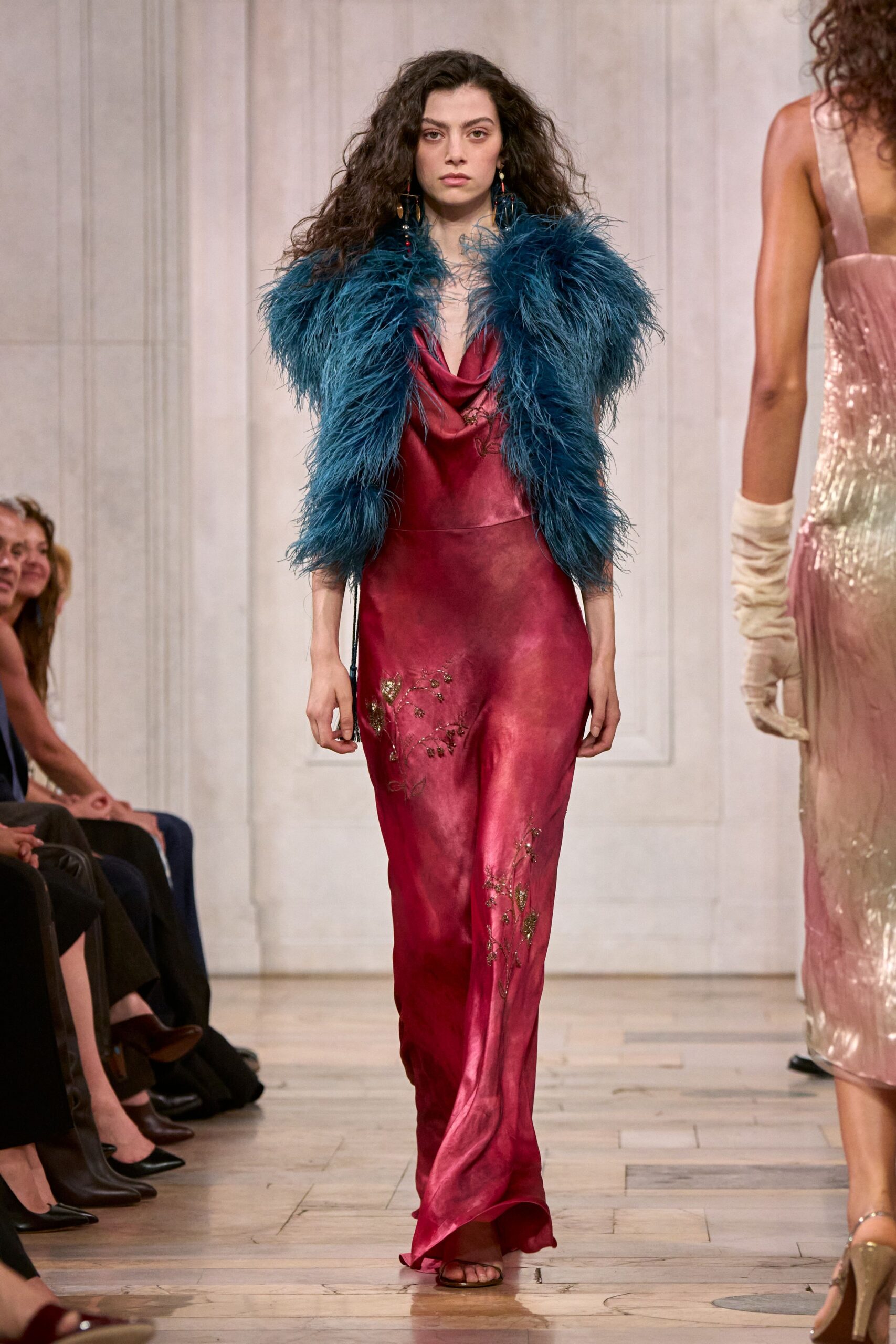 Teal feather jacket, shaggy plumes swelling past the shoulders. Magenta satin slip dress with a draped cowl and gilt vine embroidery. Long drop earrings with tiny red beads. Brown thong sandals peeking from the dragging hem. Crossing a hotel ballroom after the wedding band has packed up.