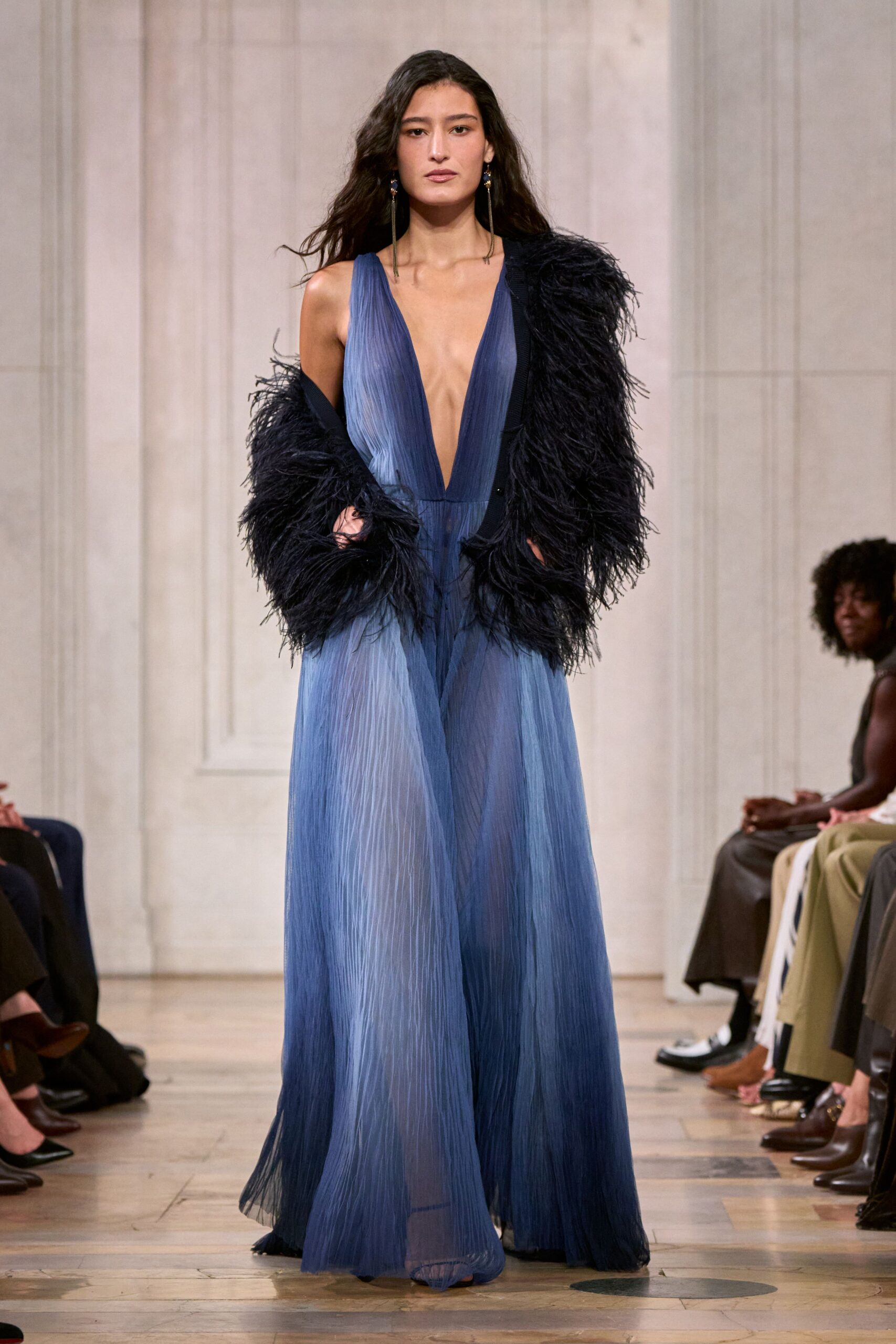 Long tassel earrings with tiny blue stones at the lobe. Plisse chiffon gown fading from slate to mist blue, plunging to the waist. Black shaggy feathered coat shrugged low off one shoulder. Crossing a hotel atrium after midnight, still carrying cold air from outside.