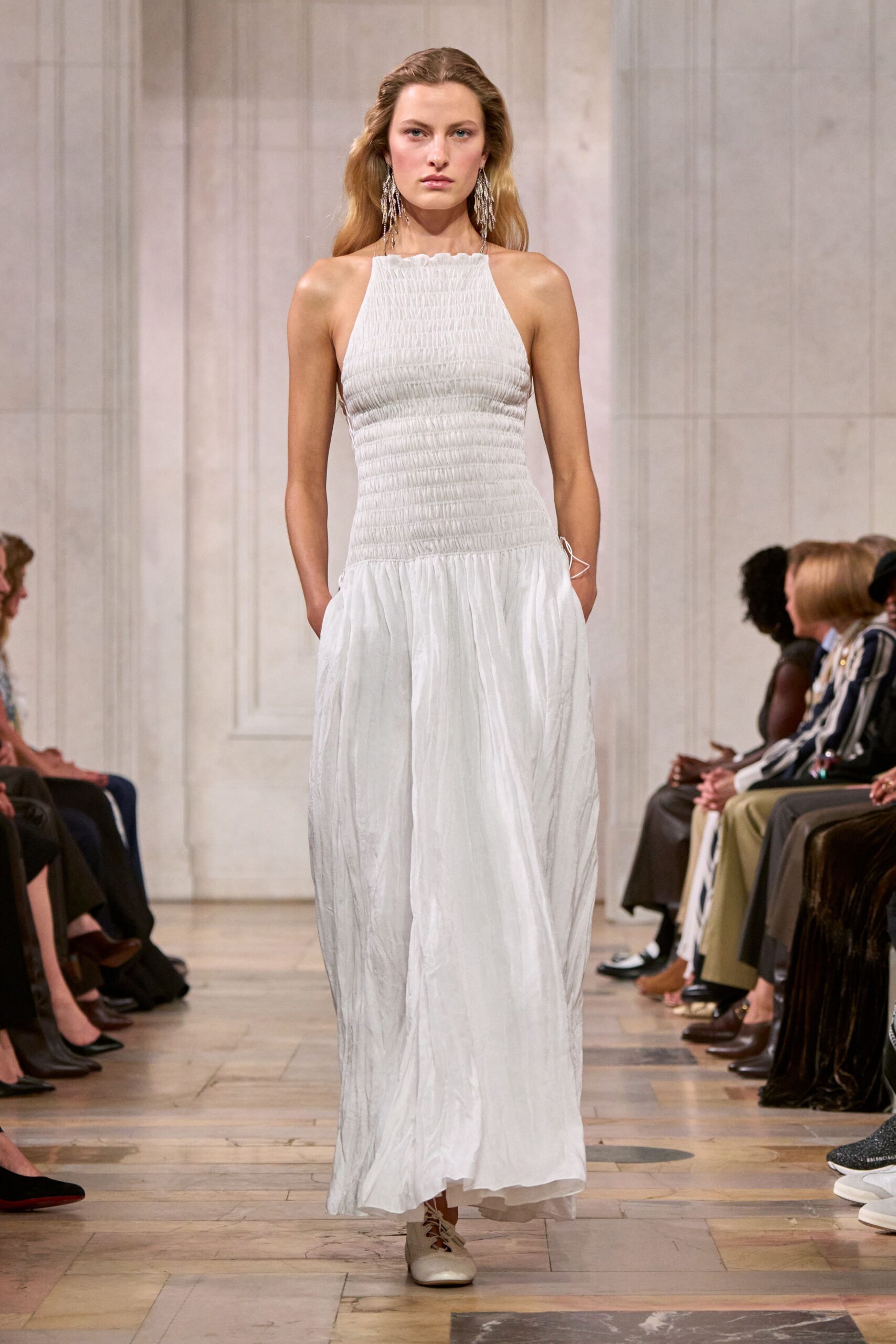 Long crystal fringe earrings brushing the shoulders. White halter maxi dress, densely shirred through the torso then falling crinkled and loose. Pale lace-up flats with a squared toe. Walking down to the jetty after a wedding, shoes sinking in the grass.