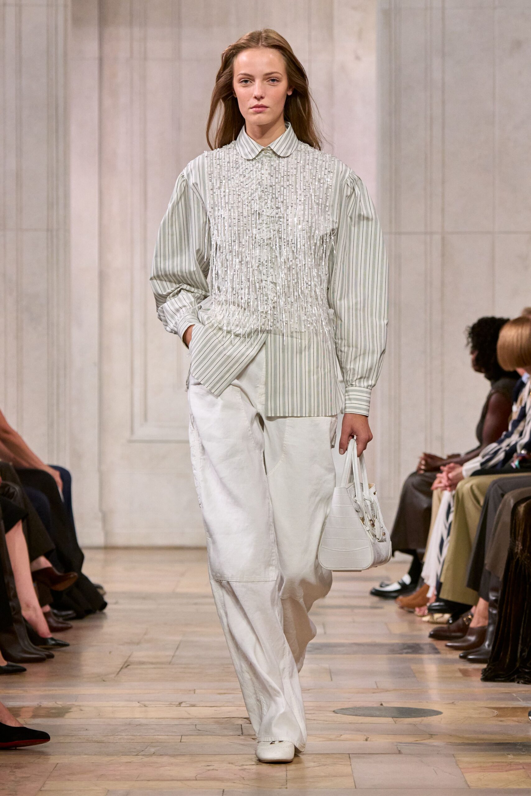 Pale sage striped cotton shirt, bib crusted with dangling clear sequins. Chalk wide-leg trousers with a low patch pocket on the thigh. White crescent handbag with crocodile embossing and tiny gold studs. Square-toe white flats. Walking into a hotel breakfast room before anyone has finished their coffee.