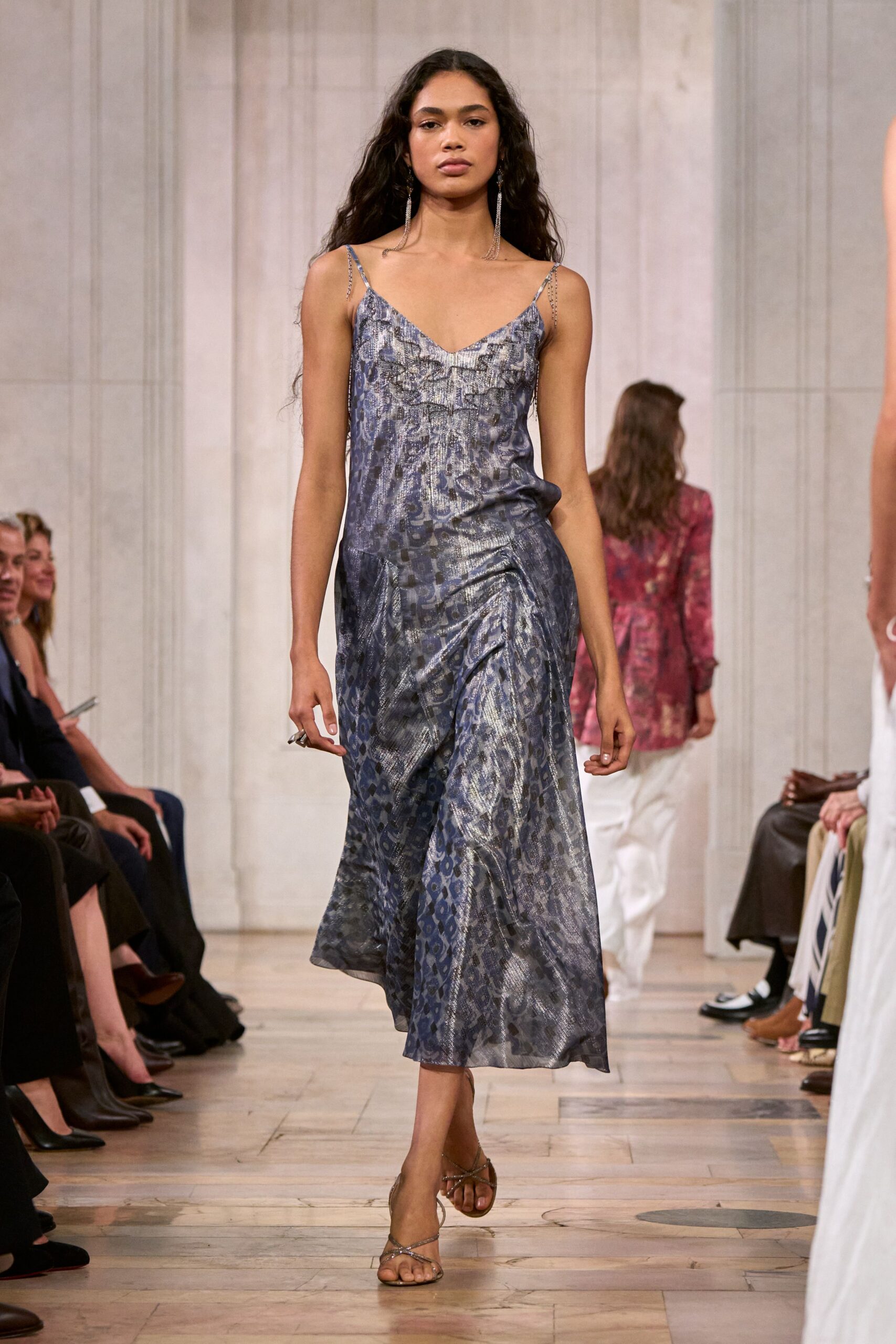 Blue-grey jacquard slip dress with silver foil shimmer and softly ruched hips. Long crystal fringe earrings brushing the collarbone. Silver thong sandals with skinny glitter straps crossing the foot. Walking out to a hotel terrace just before the heat breaks.