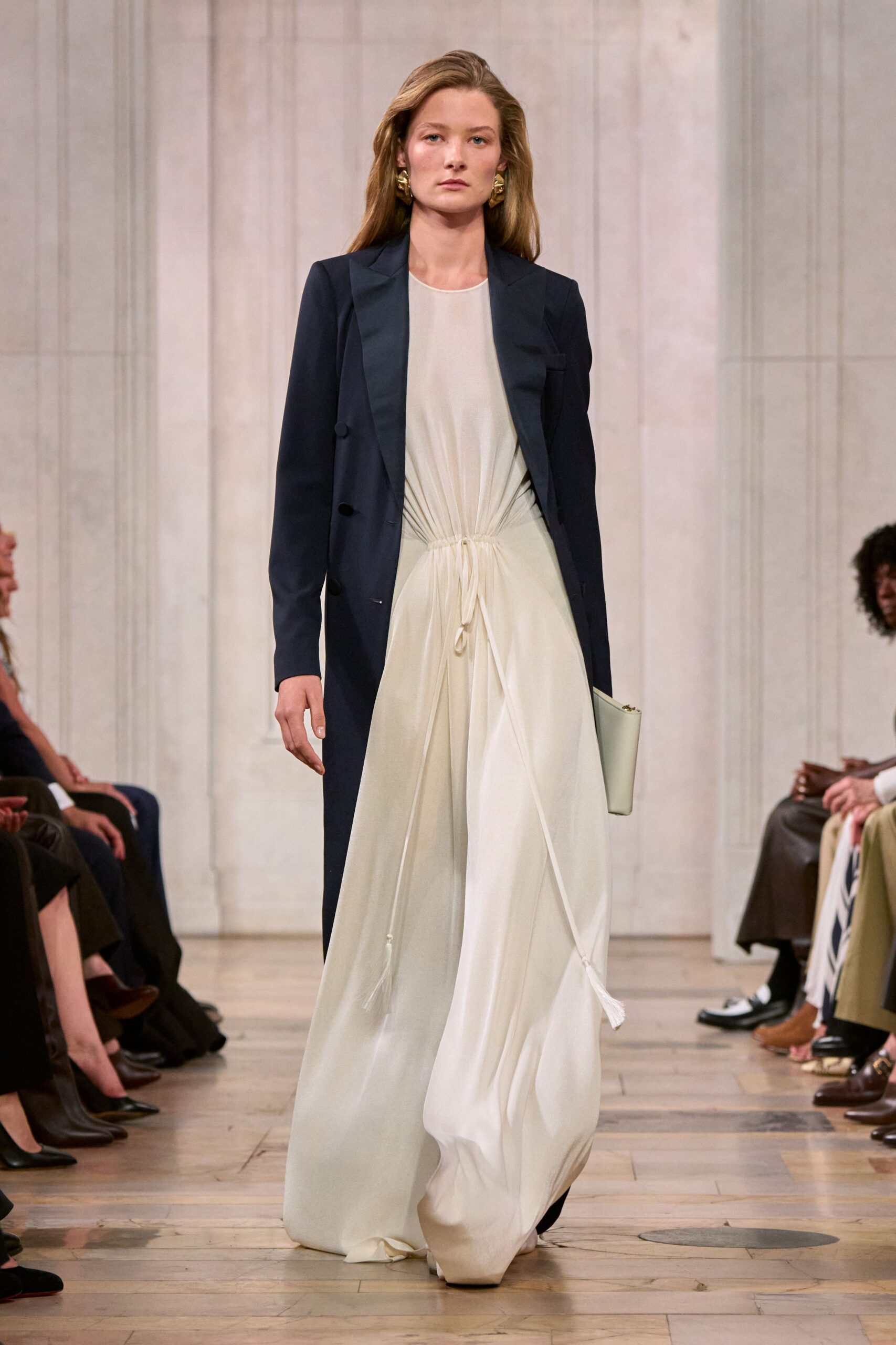 Crushed gold clip earrings, knotted like folded foil. Midnight tailored coat with satin-faced lapels and a low button stance. Ivory fluid gown, ruched to a drawstring waist with tasselled cords. Pale cylindrical clutch tucked under one arm. Crossing a museum atrium after the string quartet has packed up.