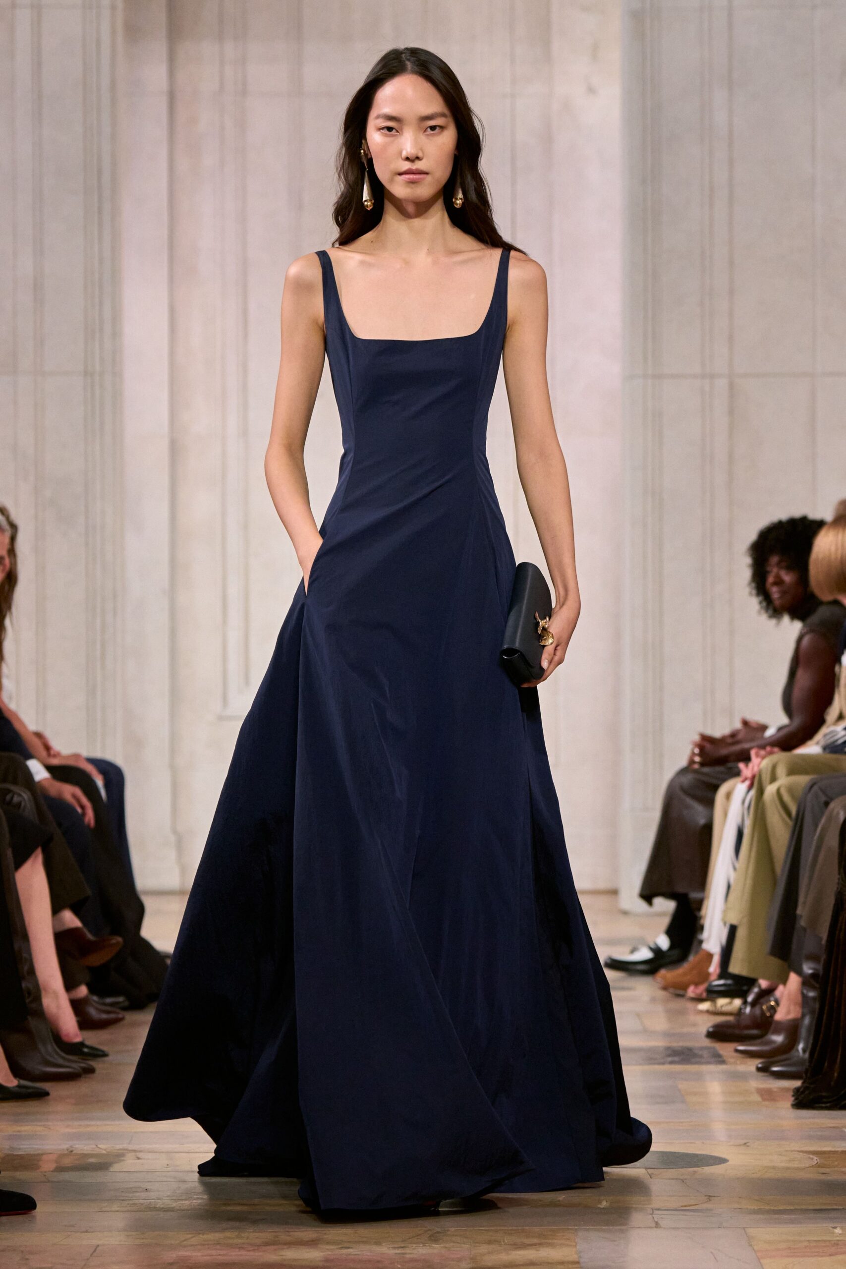 Midnight cotton gown with a square neckline and sharply seamed waist. Matte black cylindrical clutch tucked under one hand. Long gold drop earrings, one brushing the collarbone. Crossing a parquet hall after midnight, still carrying the speech notes.