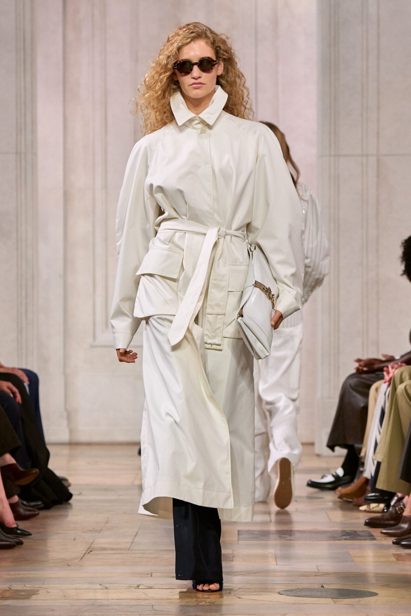 Round tortoiseshell frames with dark smoked lenses. Ivory coated-cotton coat dress, oversized collar and belt knotted off-center. White crocodile-embossed clutch tucked under the arm. Black narrow trousers breaking clean over open toes. Black mule sandals. For crossing a hotel lobby before the rain starts properly.
