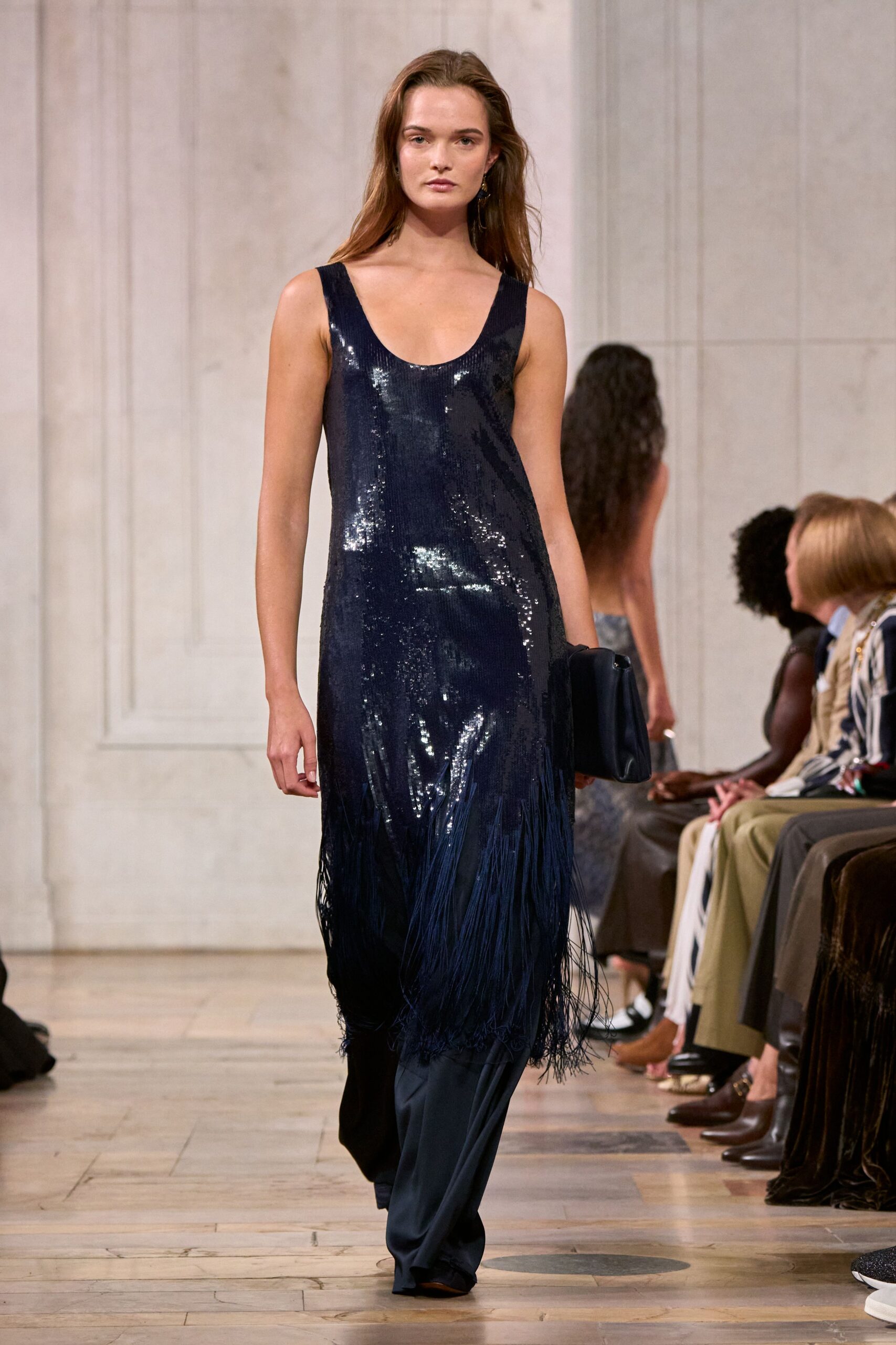Ink-dark sequined column dress with a scooped neck and swinging fringe hem. Fluid navy trousers pooling under the dress. Soft black foldover clutch tucked under one arm. Tiny drop earrings. Crossing a hotel ballroom after the band has packed up.