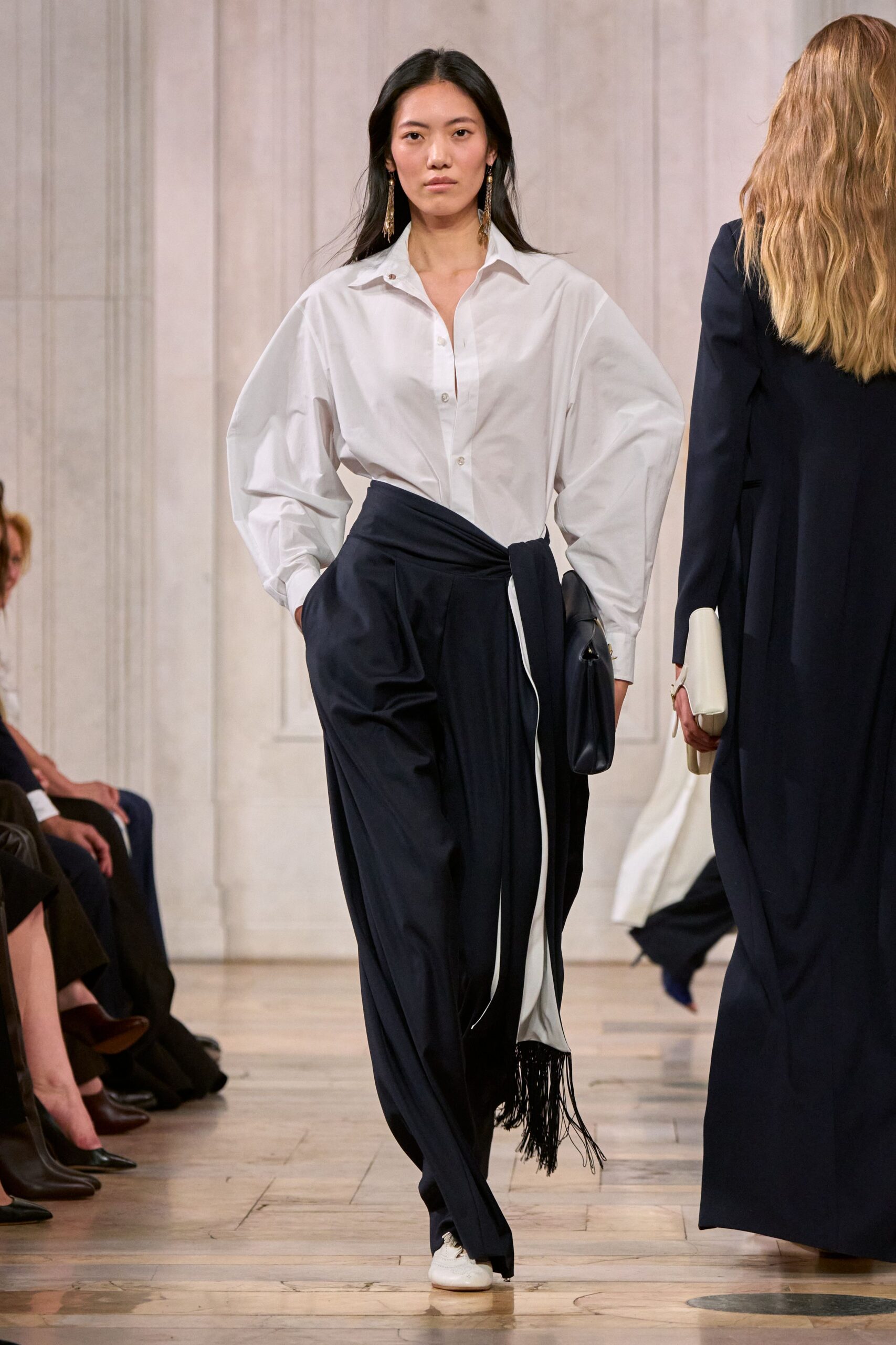 White cotton shirt with balloon sleeves and a collar spread wide. Ink-black wrap trousers, one ivory underpanel ending in swingy fringe. Soft black pouch tucked under the arm. Long gold tassel earrings. White lace-up flats with rounded toes. Crossing a parquet hall after lunch, still carrying the speech notes.