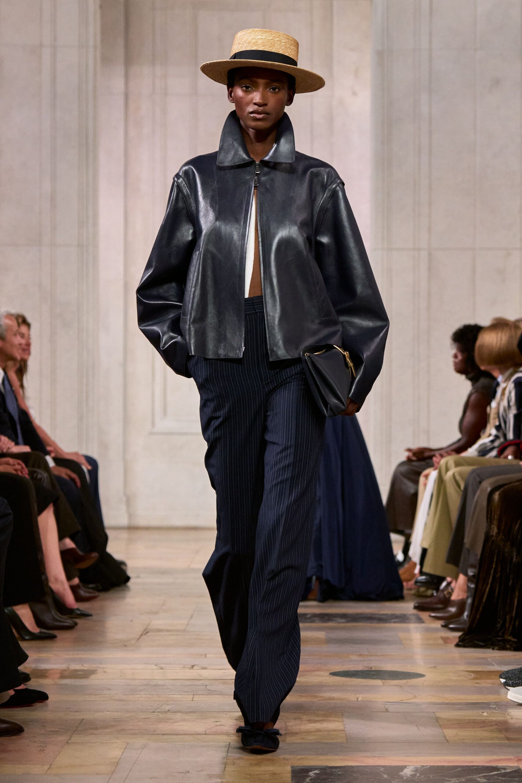 Flat-crowned straw boater with a black grosgrain band. Ink-black leather jacket, broad collar and zip left slightly split. Navy pinstriped trousers falling long over the front of the shoes. Soft black clutch with a curved gold zip. Black flats tied with tiny bows. Crossing the square after a matinee, hat still on, hands full.