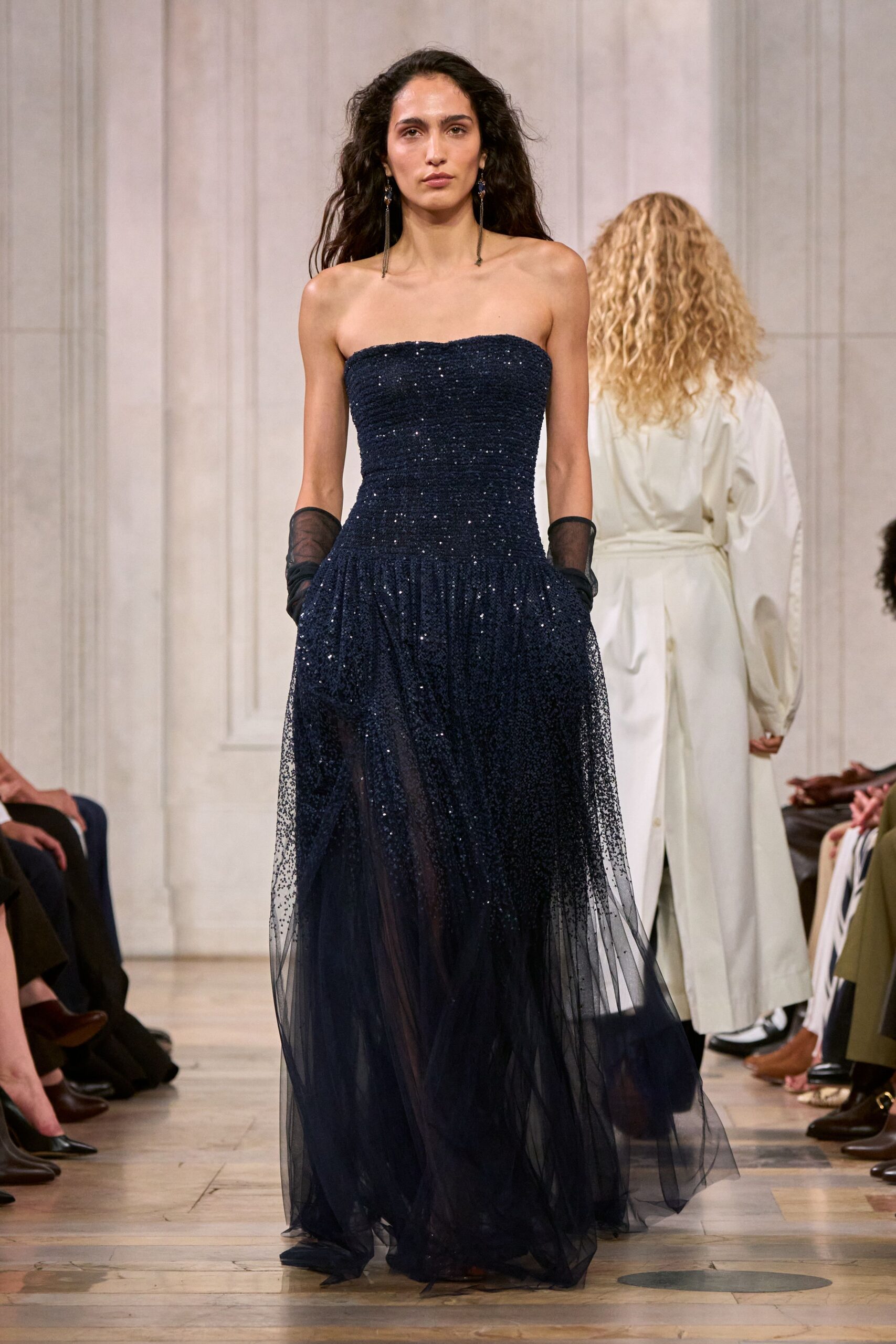 Long black tassel earrings with small cobalt drops at the lobe. Midnight strapless gown, densely sequined through the bodice then dissolving into beaded tulle. Sheer black opera gloves slouched softly below the elbow. Crossing a hotel ballroom after the orchestra has packed away.