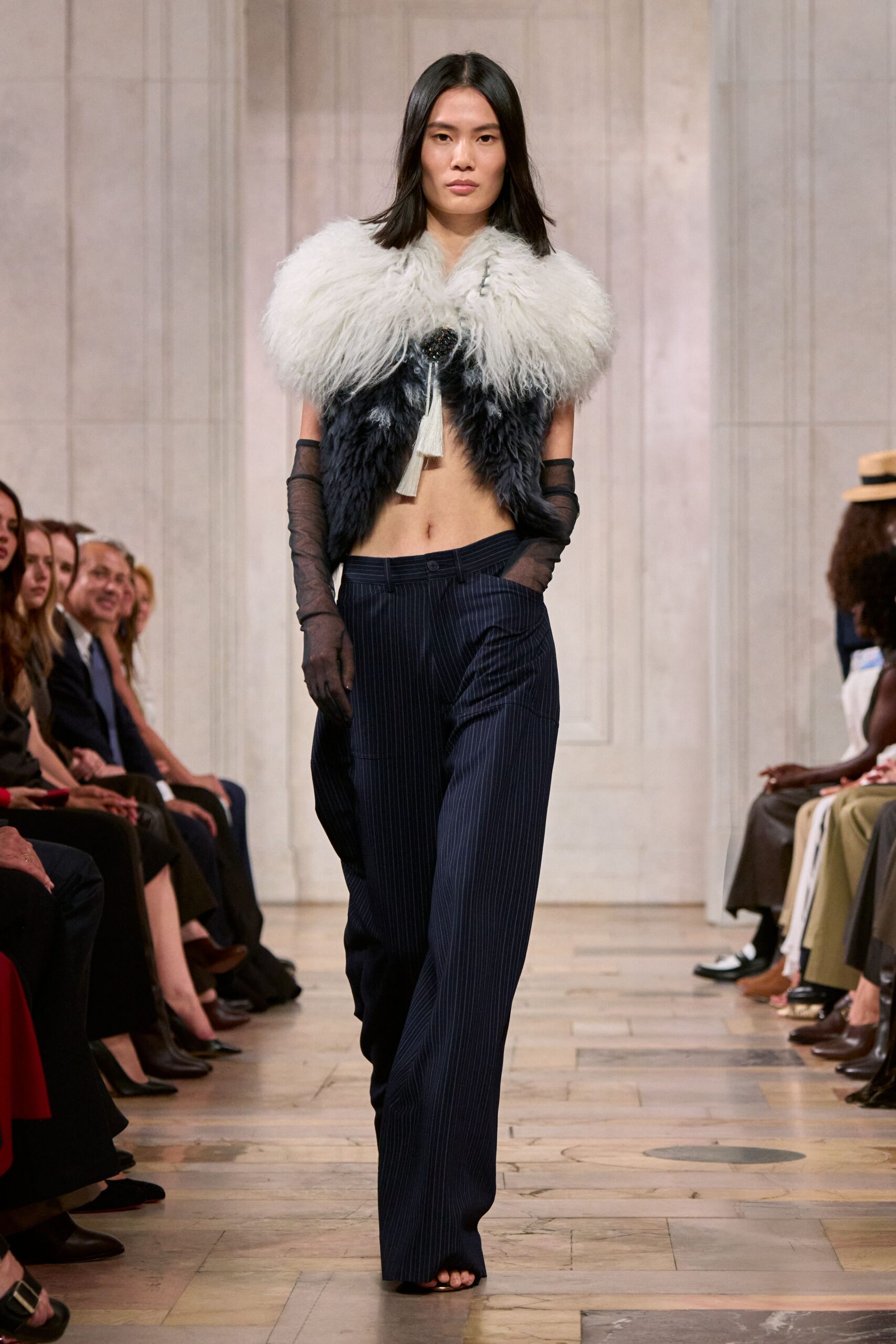 Cropped shaggy fur vest, pale at the shoulders and charcoal below. Dark jewel brooch with two cream tassels hanging at the sternum. Sheer black opera gloves, softly ruched at the forearms. Low-slung navy pinstripe trousers pooling over open toes. For crossing a hotel lobby after midnight with your shoes in hand.