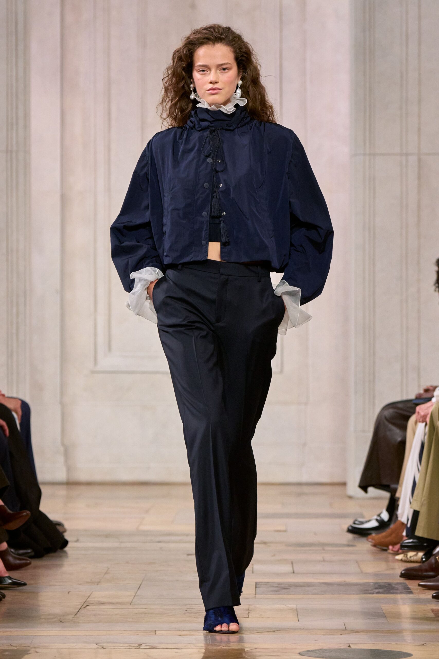 Sheer white organza ruffle collar and cuffs pushing out in soft clouds. Cropped navy shell jacket with tasselled drawcords and matte snap placket. Dark straight trousers with a clean pressed crease and narrow waistband. Blue velvet toe-band sandals. Crossing a parquet hall before the string quartet starts tuning.