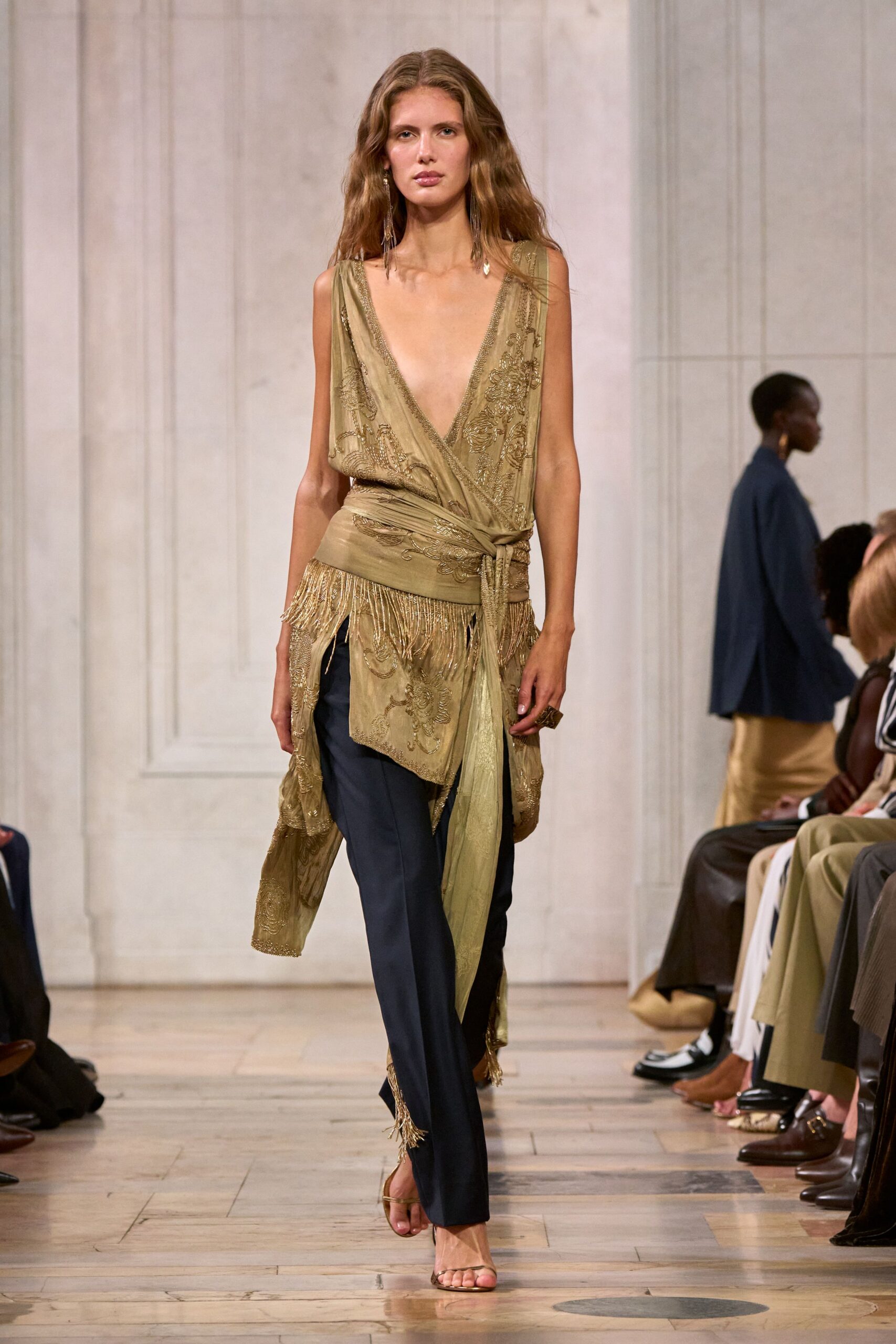 Dusty olive wrap top in sheer beaded chiffon, plunging to the waist. Wide ruched sash knotting at the hip, one tail hanging long. Ink straight trousers, fringe flicking from the outer hems. Barely there metallic thong sandals. Long chain-fringe earrings. Walking through a hotel arcade after midnight, heels ticking on stone.