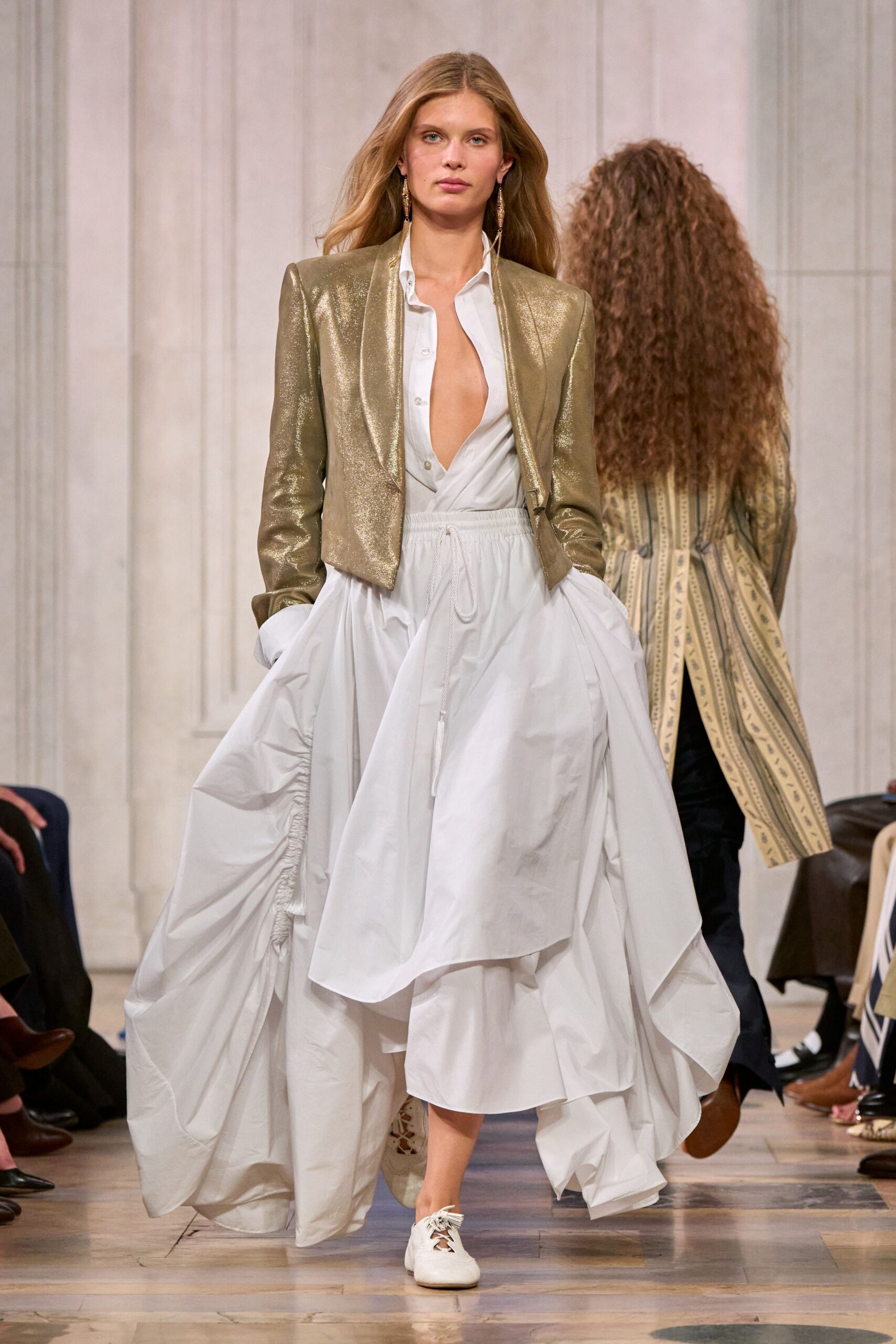 Cropped gold lame jacket with sharp shoulders and a raw-cut front. White poplin shirt dress plunging open, hem ballooned into gathered swags. Long chain earrings brushing the collar. White leather lace-ups with frayed ties. Crossing a hotel ballroom at daybreak, shoes still soft from dancing.