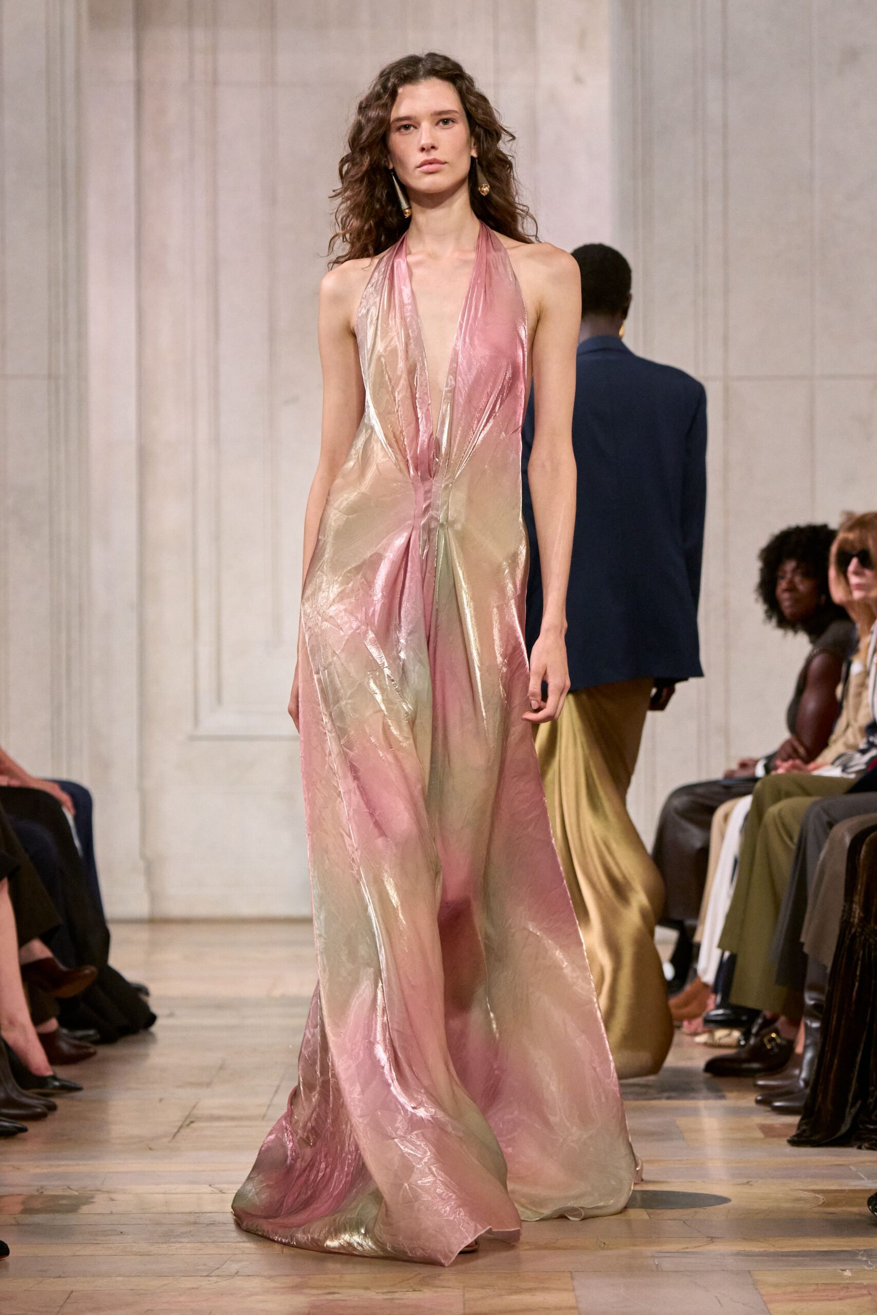 Liquid lamé halter gown in blush and pale gold, knotted low at the waist. Long drop earrings with pearl-like tips brushing the jaw. Crossing a hotel atrium after the string quartet has packed away.