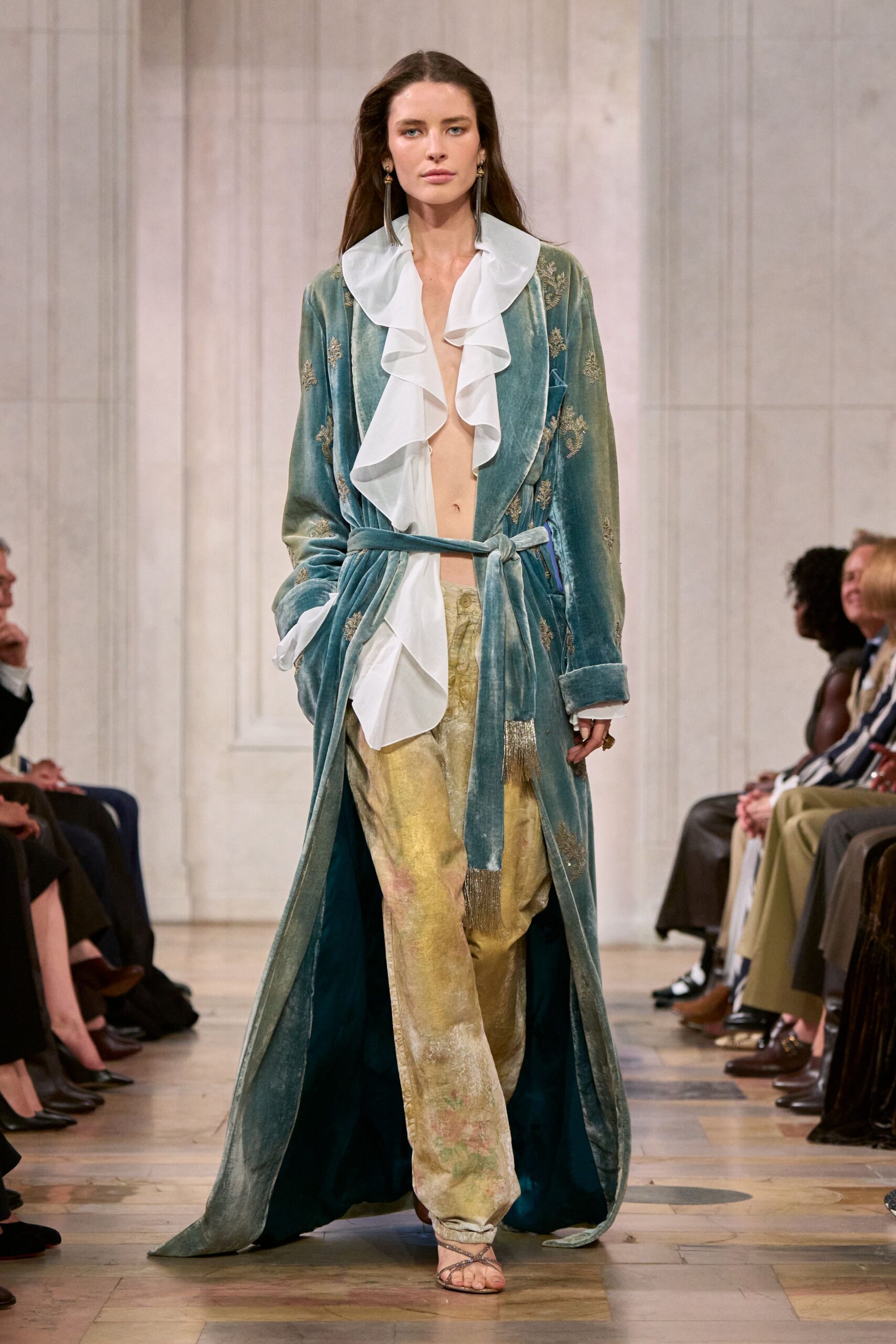 Seafoam velvet robe coat with faint metallic sprig embroidery and tasseled tie ends. White georgette blouse falling in deep loose ruffles to the waist. Pale gold crushed velvet trousers, soft floral ghosts near the shin. Silver wrap sandals. Walking through a faded seaside hotel before breakfast is served.