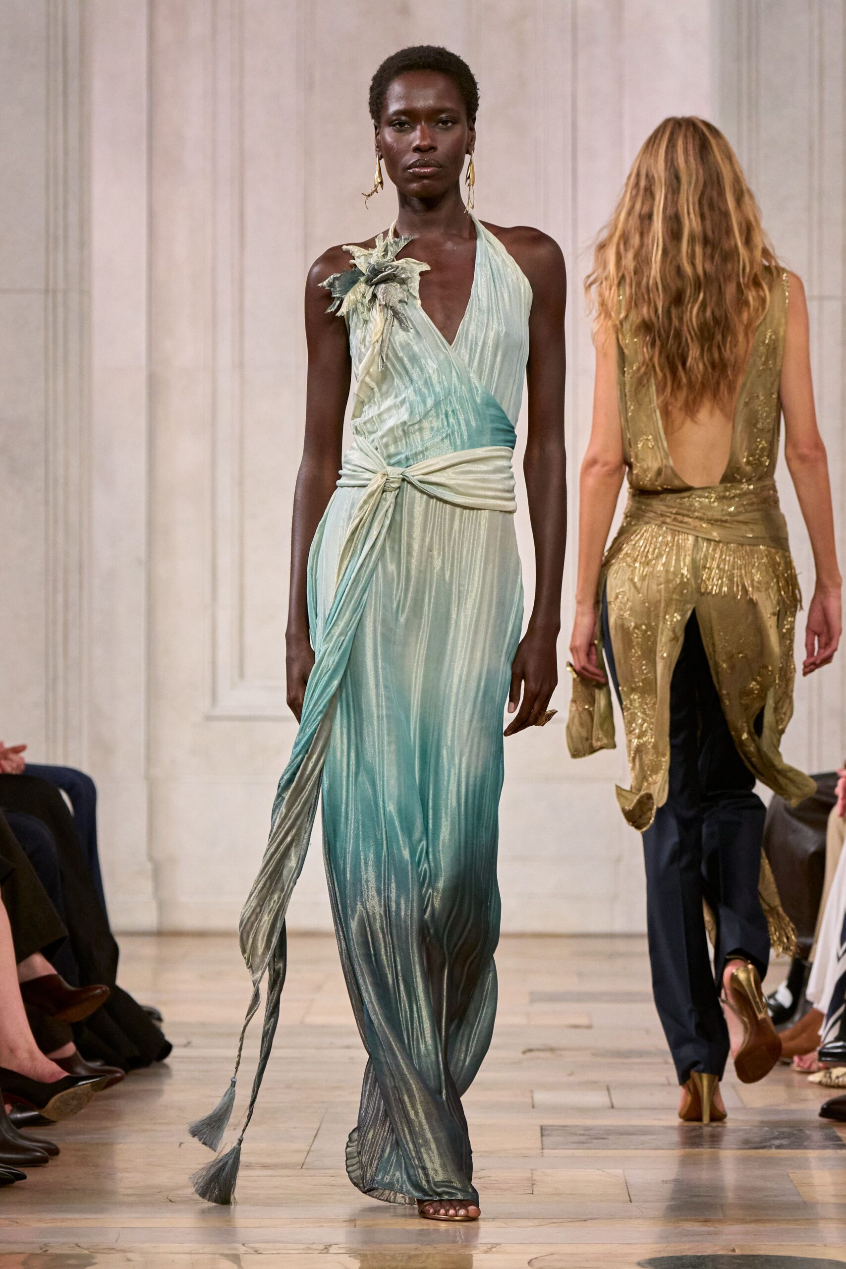 Seafoam plisse halter gown, wrapped low with a knotted sash and tassel ends. Crushed fabric flower at one shoulder, petals curling outward. Mismatched gold drop earrings. Large sculptural ring on one hand. Crossing a hotel terrace just before the storm reaches the bay.