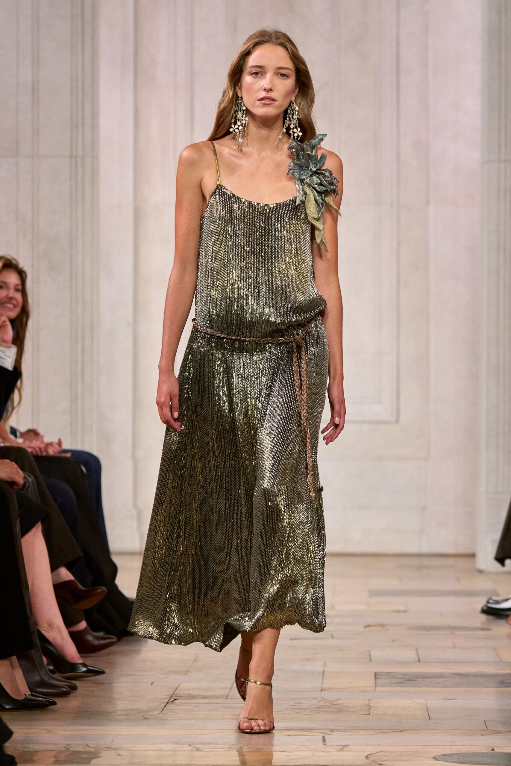 Antique-gold chainmail slip dress, bloused at the waist with a liquid drape. Oversized pewter fabric flower pinned hard onto one strap. Braided cord belt, one end hanging nearly to the calf. Long crystal cluster earrings. Thin metallic ankle-strap sandals. Hotel ballroom after midnight, collecting wilted stems from the tables.