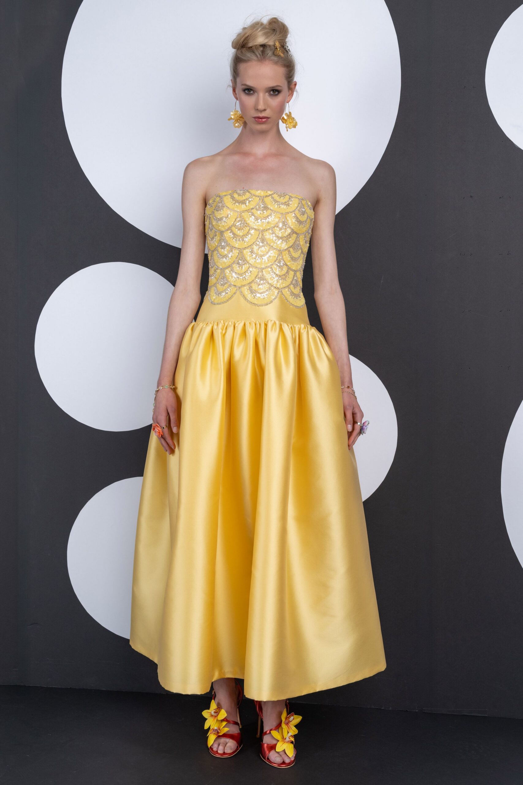Strapless butter-yellow satin gown, silver beaded scallops over a dropped gathered skirt. Yellow flower cluster drops. Red satin stilettos with oversized yellow orchid corsages. Garden wedding, cake already cut and the grass starting to stain hems.