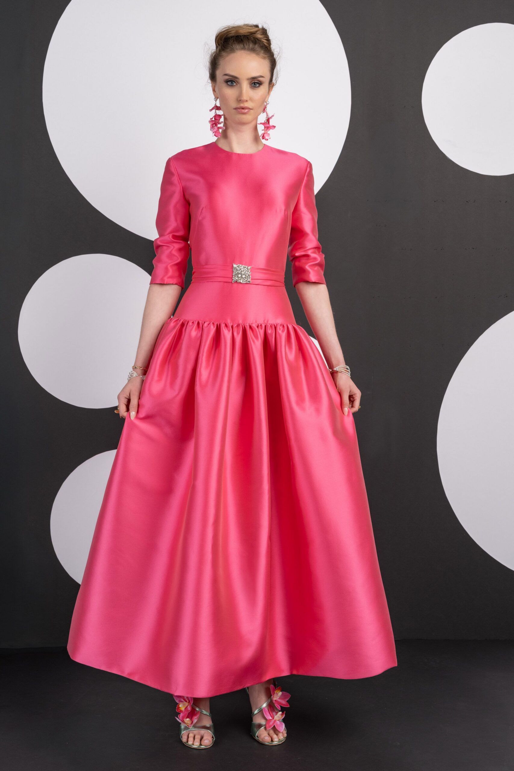 Hot-pink satin dress with bracelet sleeves, dropped gathered waist and crystal buckle. Cascading pink orchid drops brushing the jaw. Metallic ankle-strap sandals with orchid blooms over the toes. For cutting the cake in a marquee while the grass is still damp.